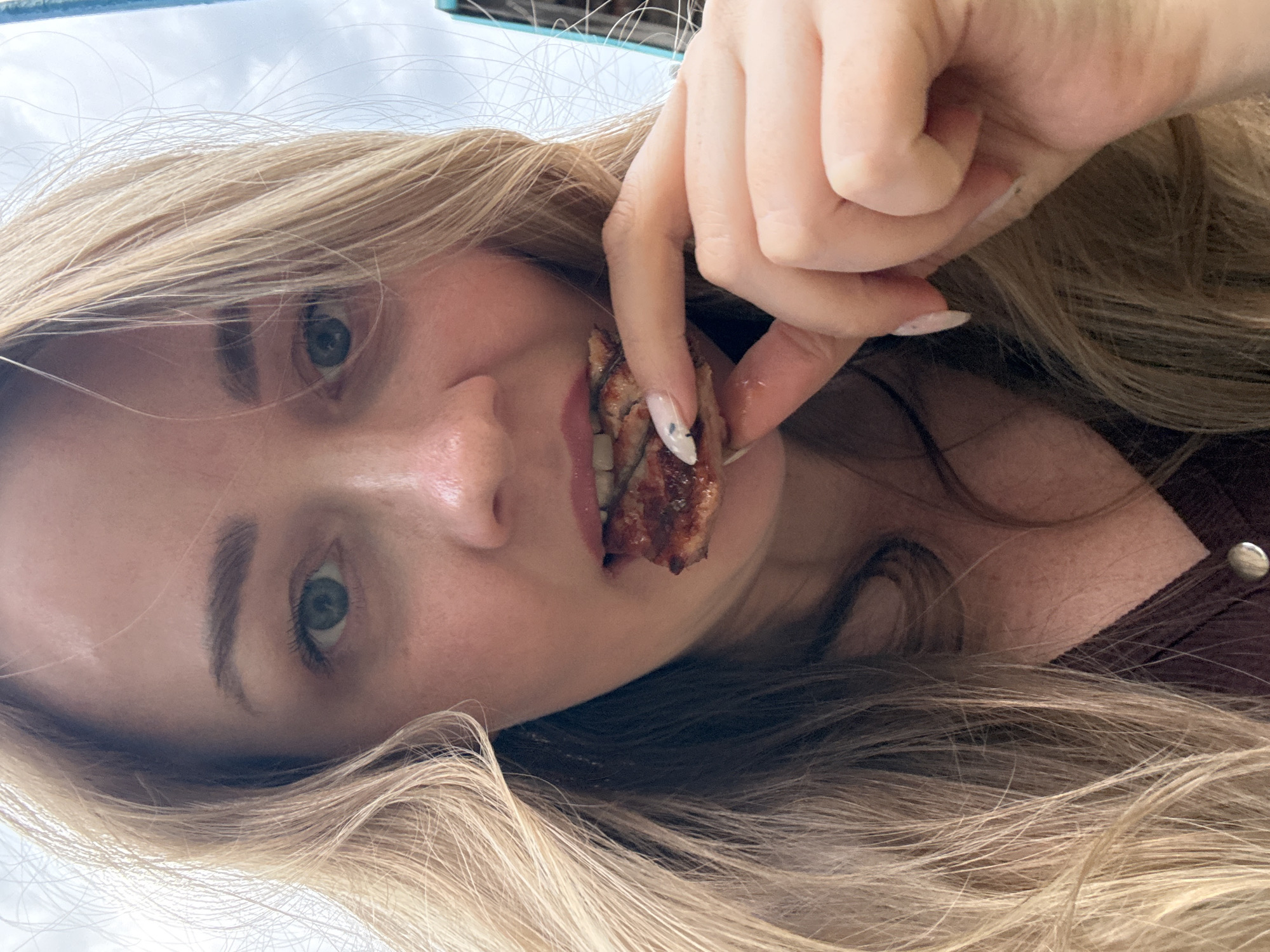 A young woman with long, blonde hair is holding a piece of cooked meat to her mouth. She has green eyes, fair skin, and painted nails. She is wearing a brown top. The background is blurry and shows a bright, cloudy sky. The city of Chaikovsky, Russia is not discernible in the image.