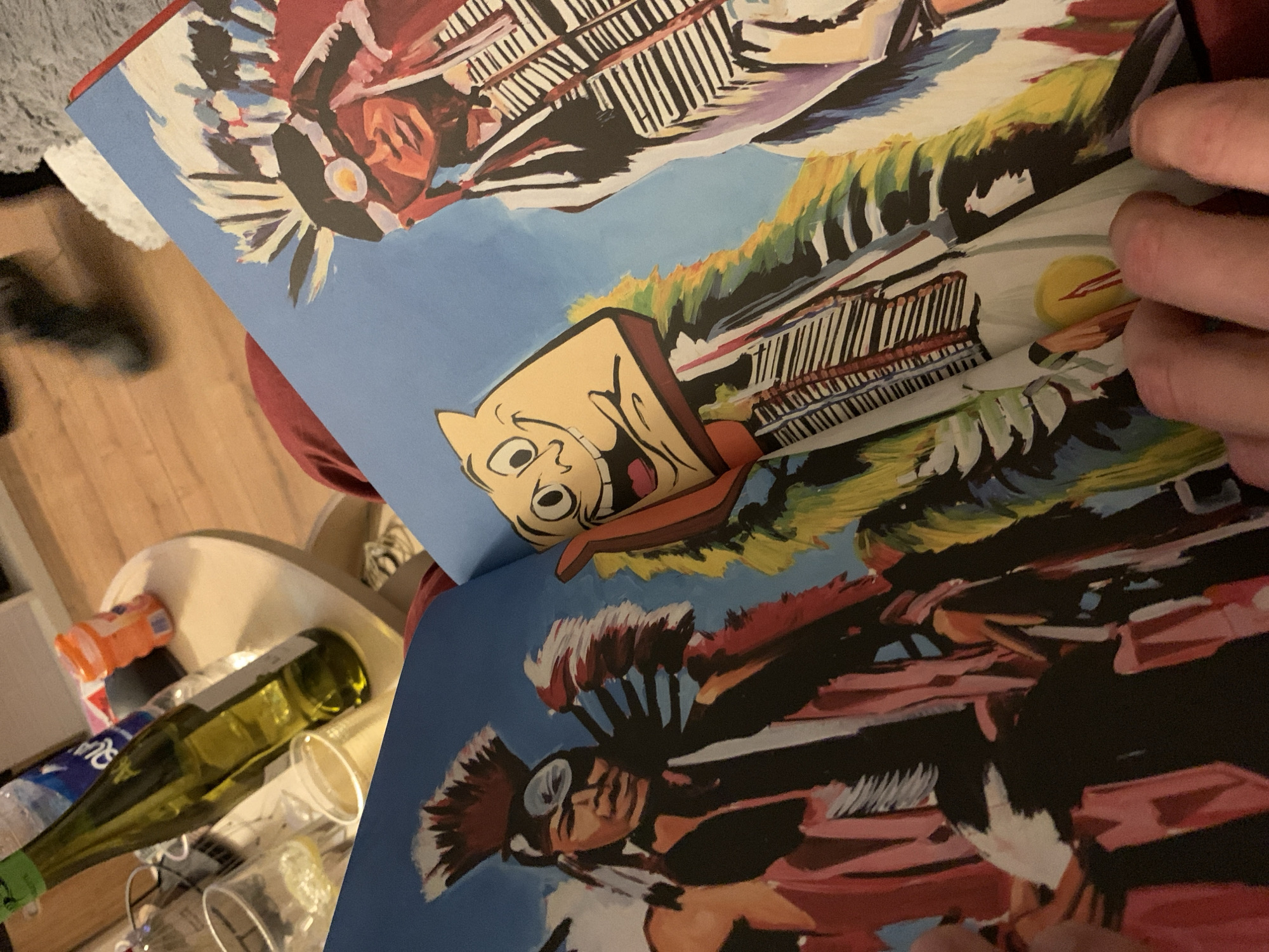 The image shows an open book with illustrations. The pages feature stylized illustrations with figures resembling Native American characters and a cartoon-like face. One illustration includes a depiction of what appears to be architecture or structures with horizontal lines and patterns.
The scene is situated in a domestic indoor setting. In the background is a cluttered area, including a green wine bottle, plastic cups, and other miscellaneous items on a table.
There are no identifiable individuals in Tel-Aviv, Israel.