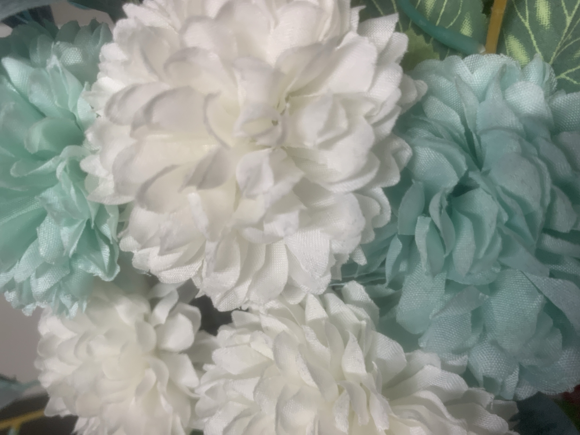 The image is a close-up, overhead shot of several artificial flowers, predominantly white and a light teal color. The flowers have numerous layered petals, giving them a fluffy appearance. Some of the teal flowers have green leaves and stems visible around them. The background is blurred and dark. There are no people or animals present, and no discernible environmental context related to the city of Sinyavino, Russia, or any specific location. The scene appears to be a still-life arrangement of decorative items.
