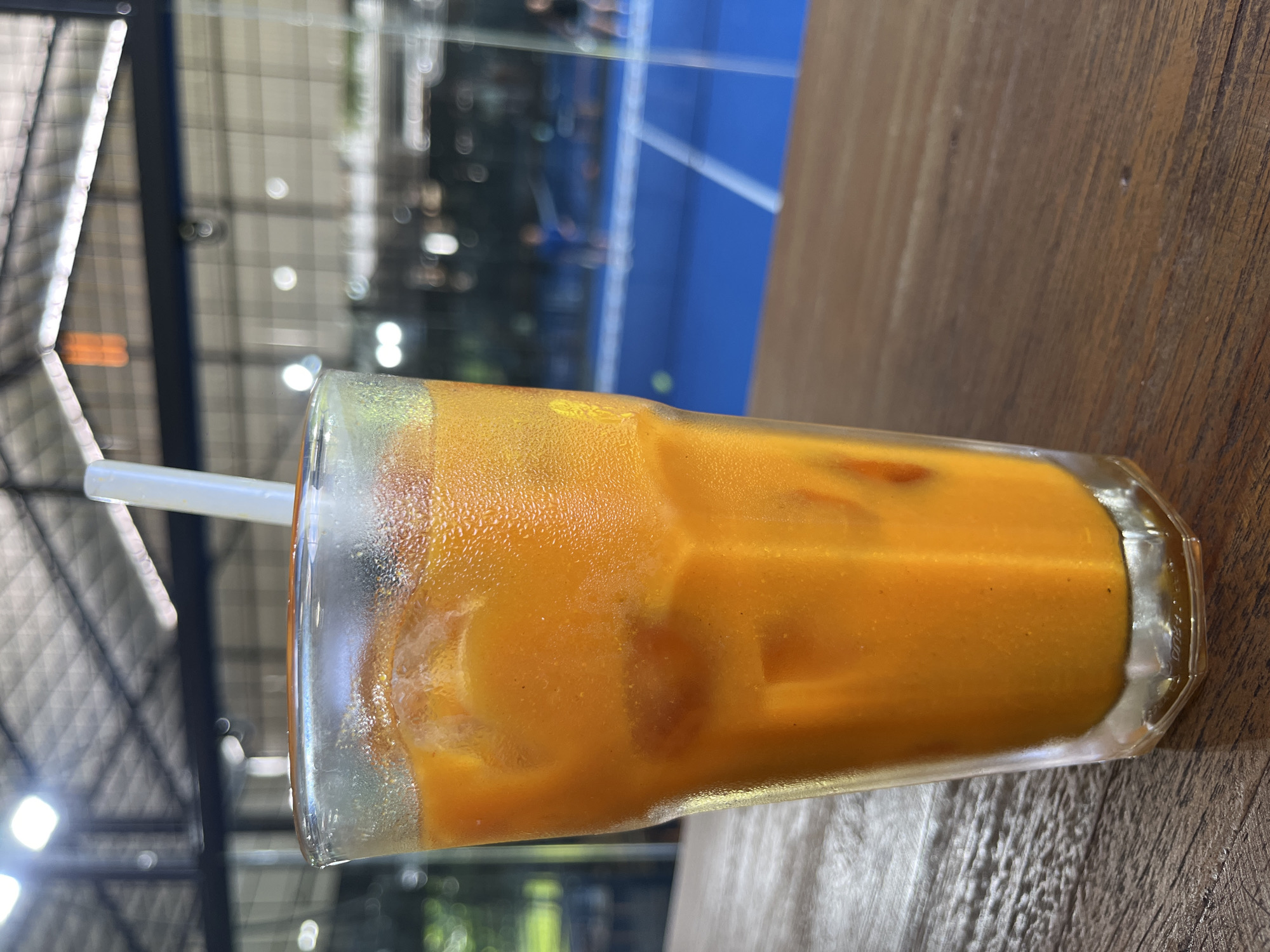 The image displays a tall glass of an orange-hued iced drink, likely a Thai iced tea given its distinct color and visible sediment, resting on a dark brown wooden table. The glass is clear and heavily condensed on the outside, indicating a cold beverage in a warm environment. Numerous ice cubes are visible within the liquid, and a white plastic straw is inserted into the drink.
The scene is set outdoors or under a covered area, with a softly blurred background. A prominent bright blue line, characteristic of a court boundary (such as for tennis or padel), is visible in the distance, suggesting the location is adjacent to a sports facility. Above this, a metal framework or netting appears to be part of a structural roof or enclosure, with several bokeh light sources scattered throughout the background. The bright lighting suggests it is daytime.
The image captures a moment of refreshment, possibly during a break from or observation of sports activities in North Kuta, Indonesia. No visible text is present in the media.