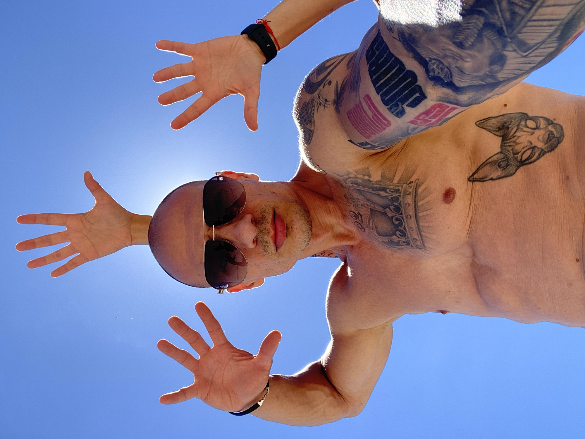 This is a low-angle, full-body shot of a bald, tattooed man standing outdoors against a clear blue sky. He is wearing sunglasses and has his arms raised, with his hands open, reaching upwards. The man's chest and arms are visible and heavily tattooed. One tattoo on his chest depicts a crown, and another on his abdomen shows a Sphinx cat's head. His left arm has a tattoo with what appears to be an American flag design and some text. He is also wearing a black bracelet on his left wrist and a black fitness tracker with a red string on his right wrist. The bright sunlight casts a lens flare on the left side of the image, obscuring a portion of the sky. There is no indication of the specific location in Pleven, Bulgaria.