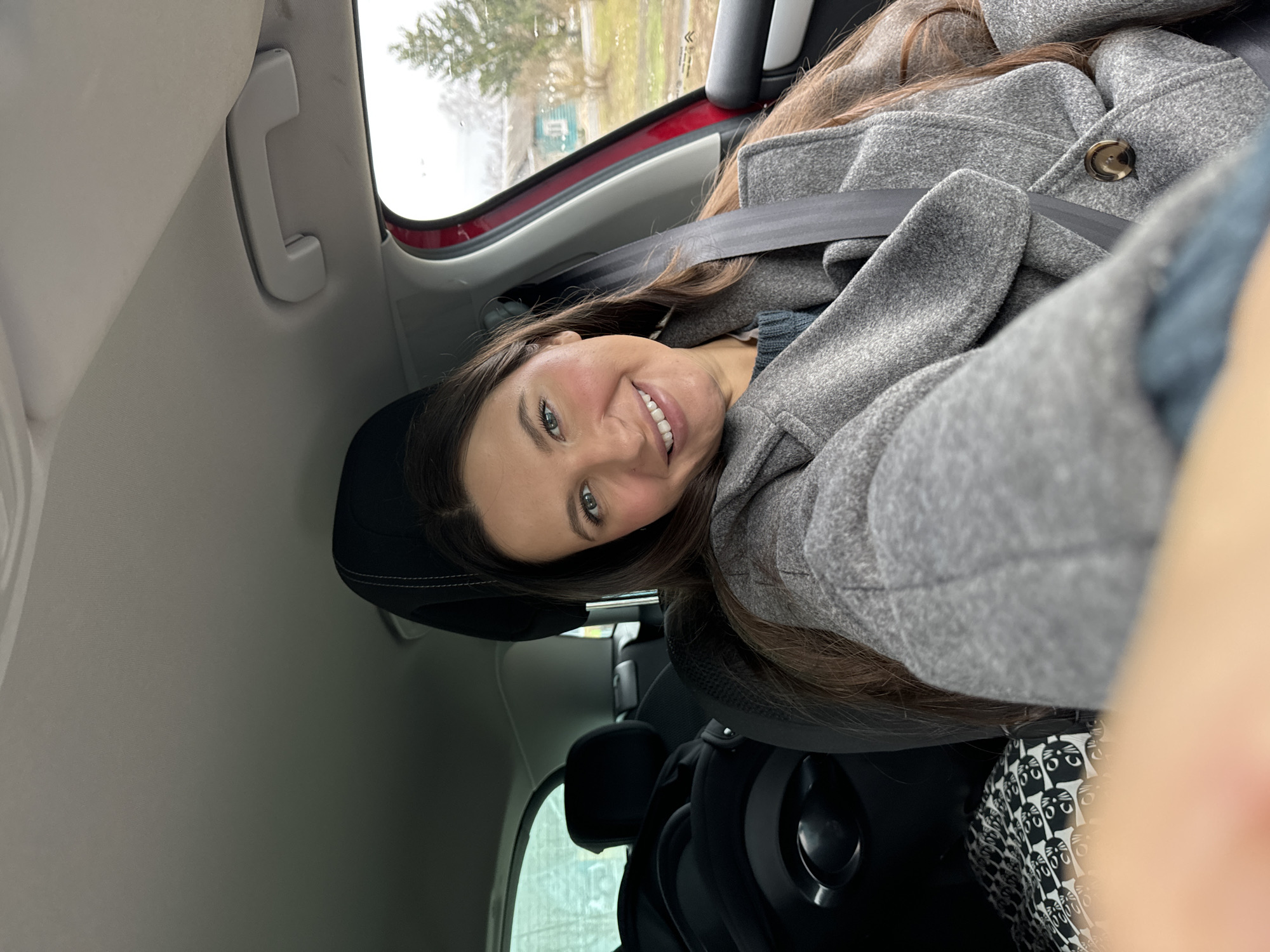 A woman wearing a gray coat and a seatbelt is sitting in a car. Outside the car window is a view of a tree and a building in what appears to be Красный Маяк, Russia. The woman is smiling and looking at the camera.