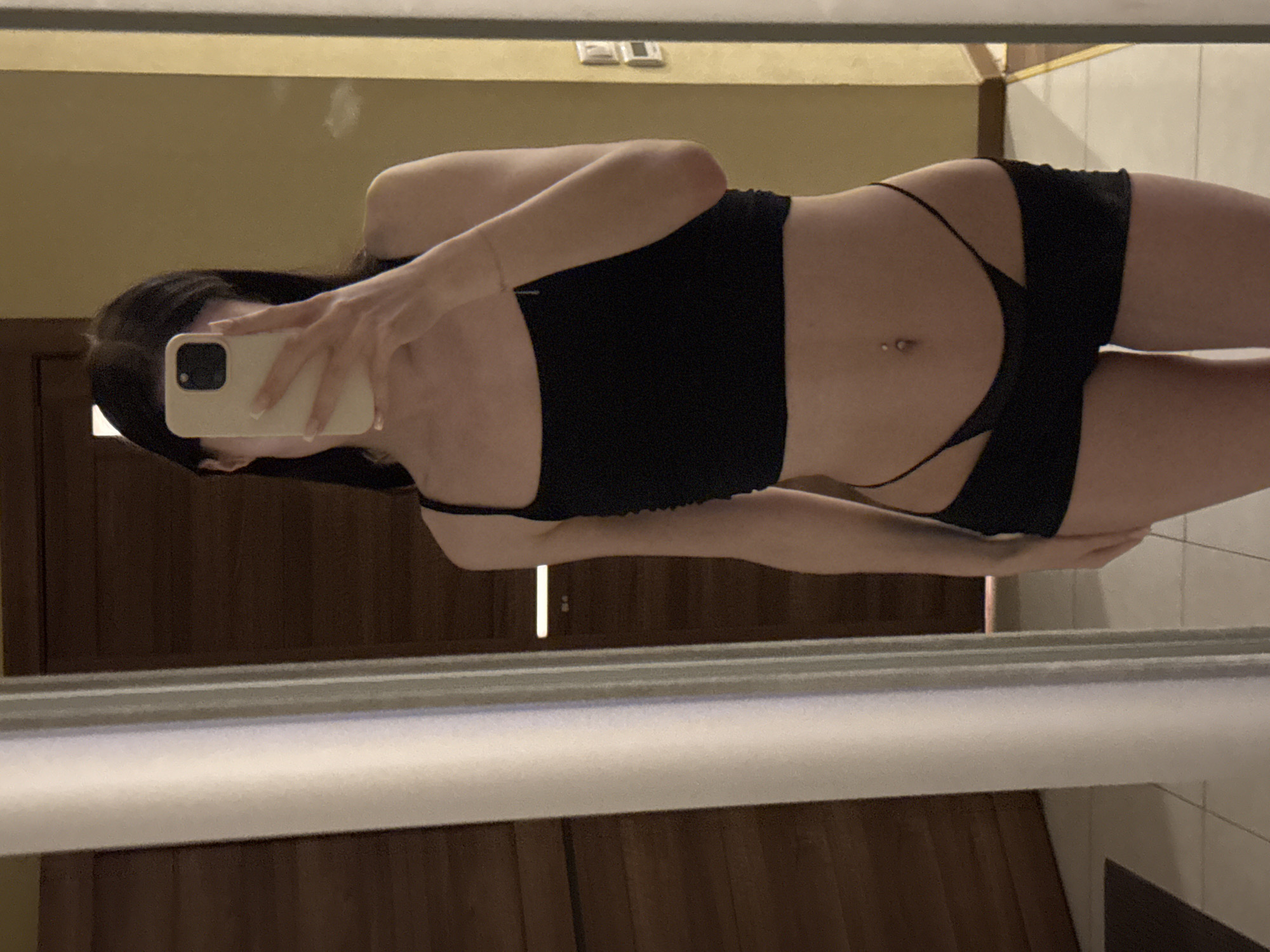 In Khimki, Russia, a young woman stands in front of a mirror, taking a selfie. She is wearing a black crop top and black shorts. Her phone is held up to her face, obscuring it from view. The background appears to be a room with wooden doors and tiled flooring. The lighting is dim, creating a somewhat intimate atmosphere. The reflection in the mirror shows the woman's full body, highlighting her attire and the surrounding environment.