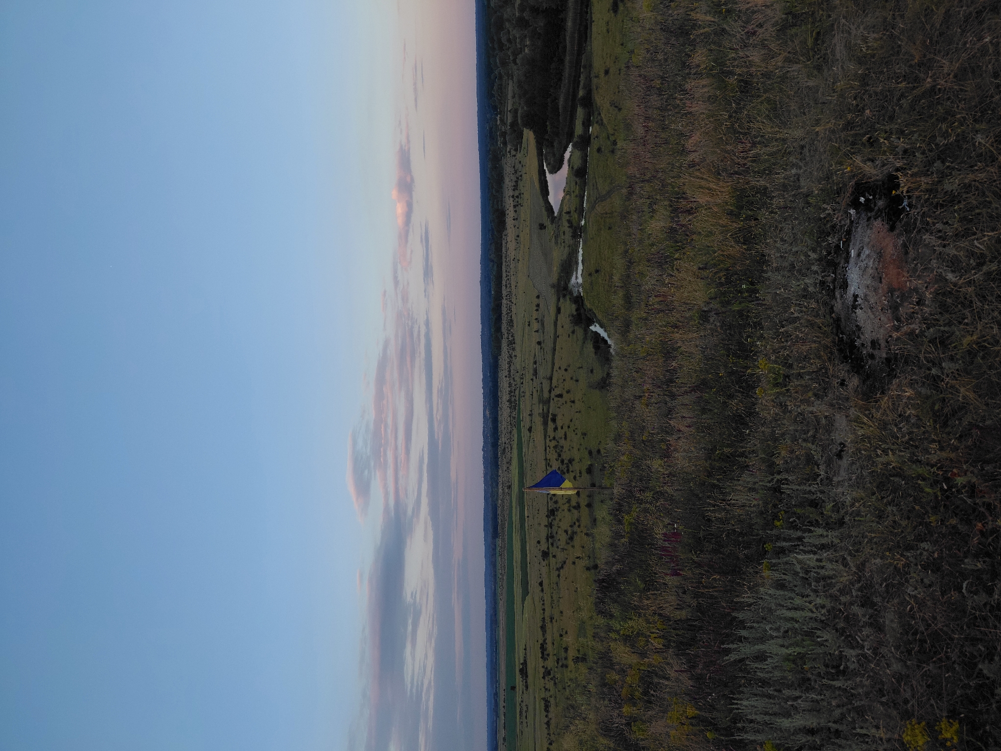 A panoramic view of a rural landscape at dusk. The sky is a gradient of pale blue to soft pink, with scattered clouds. In the foreground, tall, dry grass covers a hillside. A Ukrainian flag, blue and yellow, stands on a wooden pole in the middle ground, to the left of a winding river. The river reflects the fading light of the sky. Fields and trees stretch to the horizon. The scene appears to be located in the vicinity of Ustyvytsia, Ukraine, a village in the Sumy Oblast.