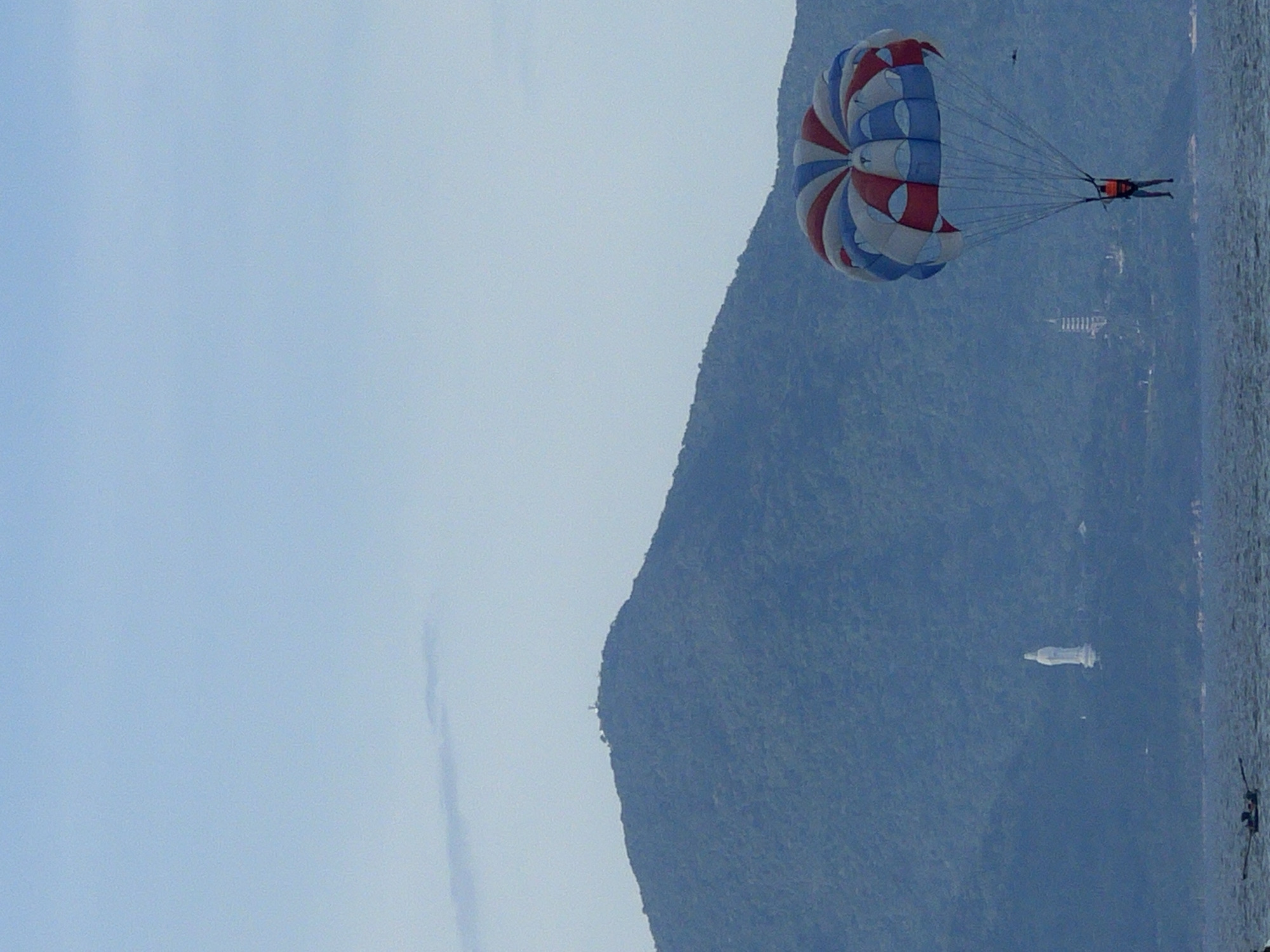 This image captures a parasailing scene near the Son Tra Peninsula in Da Nang, Vietnam. A person in an orange vest is suspended beneath a red, white, and blue parachute. The parachute is high in the sky, against a pale blue, hazy sky. In the background, the lush green slopes of the Son Tra Peninsula dominate the scene. A prominent white statue of a deity, likely the Lady Buddha of Da Nang, is visible on the hillside. Further in the distance, a slender, modern tower structure, possibly part of the Linh Ung Pagoda complex, can be seen. The foreground shows the shimmering surface of the sea, with ripples indicating a gentle breeze. A small boat with two individuals is visible in the lower left corner, and another boat is further to the right on the horizon. The overall impression is a serene coastal landscape with recreational activity.