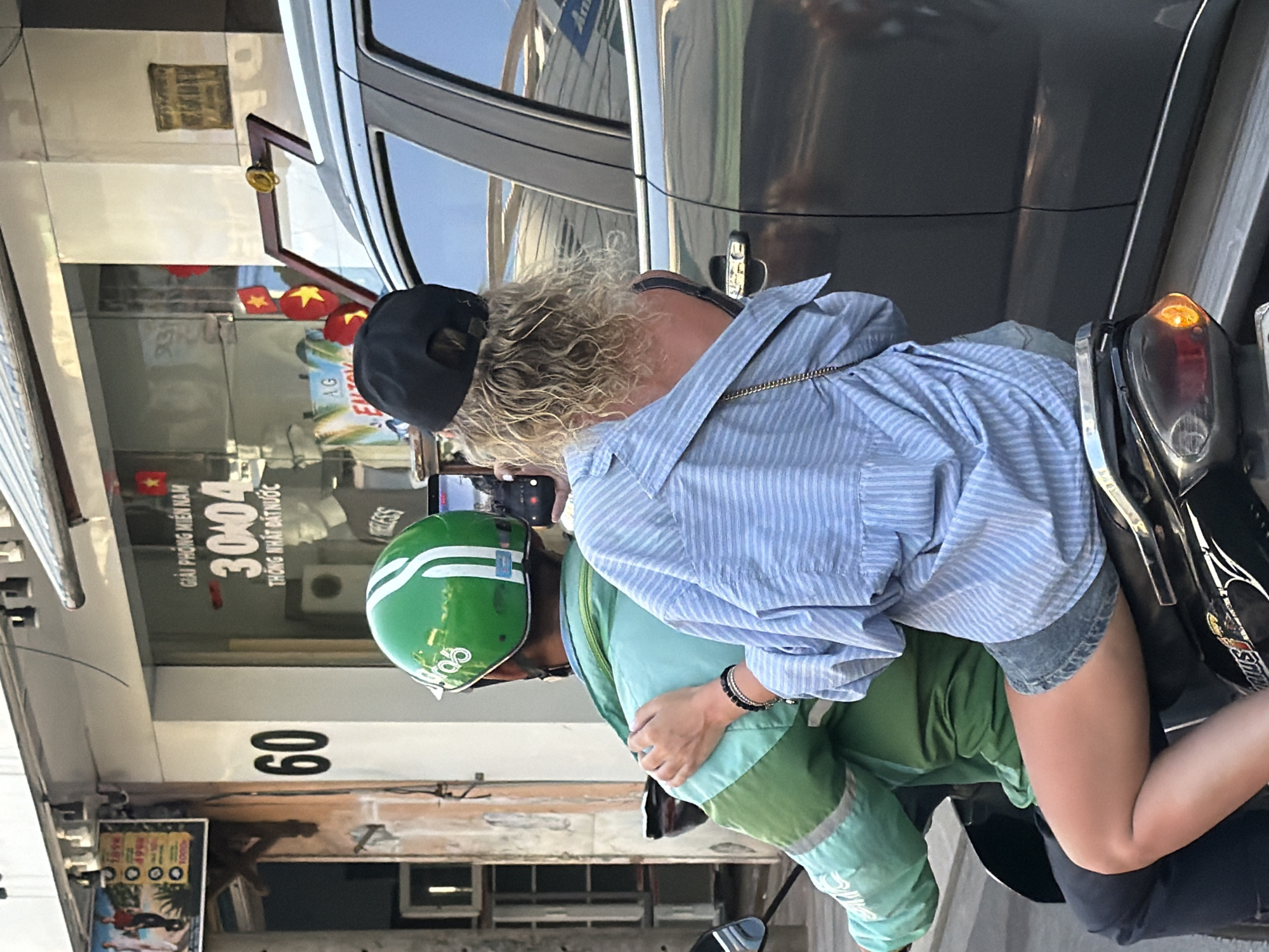 A woman with curly blonde hair wearing a black baseball cap and a light blue striped button-up shirt over a dark-colored tank top sits on the back of a motorcycle. She is holding a smartphone in her right hand, seemingly taking a picture or video. Her left arm is around the shoulders of the motorcycle driver, who is wearing a green helmet with white stripes and a teal and green jacket. The motorcycle is moving on a street in Nha Trang, Vietnam, next to a dark-colored car. In the background, a storefront displays Vietnamese text, including "GIẢI PHÓNG MIỀN NAM" and "30/04 THỐNG NHẤT ĐẤT NƯỚC," indicating a historical reference to the reunification of Vietnam on April 30, 1975. The street appears to be in a commercial area with building facades and signs visible.
