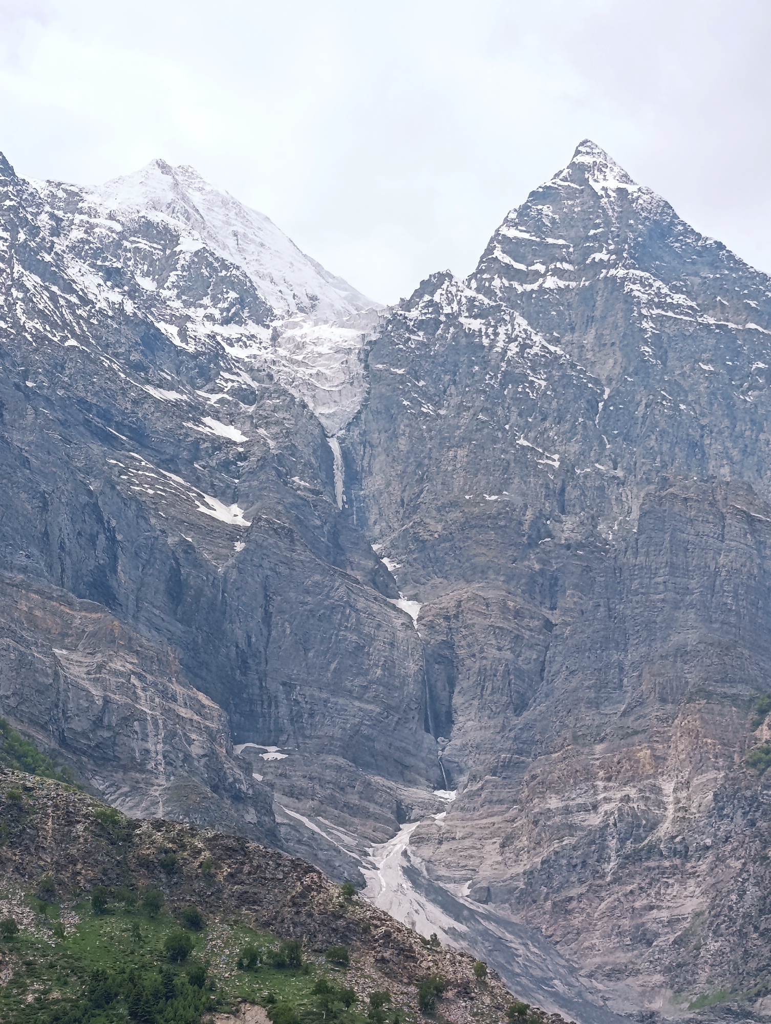 This is a vertical, medium shot of a majestic mountain landscape under a cloudy sky. The dominant subjects are two prominent, snow-capped mountain peaks that rise steeply. The left peak is broader and more rounded at its summit, while the right peak is sharper and more pointed. A glacier or large snowfield descends from the saddle between the peaks, feeding a visible waterfall that cascades down the rugged, rocky mountainside.
The mountains themselves are characterized by steep, textured slopes, showing layers of rock in shades of gray and brown. Patches of snow and ice are scattered across the rock faces, particularly at higher elevations. In the foreground, the lower slopes of the mountains are covered in dense green vegetation, including trees and shrubs, suggesting a lower altitude or a transition zone. The overall scene conveys a sense of immense natural beauty, ruggedness, and the power of geological forces. The weather appears overcast, contributing to a slightly muted color palette but highlighting the dramatic topography. The time of day is not definitively discernible due to the cloud cover. The location cues point to Jagle, India, a region known for its mountainous terrain. There is no visible text in the image.