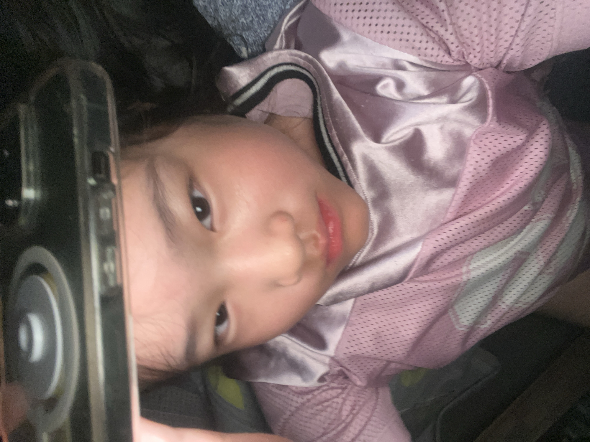 A young child is lying down, holding a smartphone in front of their face. The child has dark hair, brown eyes, and pink cheeks. The child is wearing a pink and white athletic-style jersey. The background is dark and indistinct. The location is Unknown, Unknown.