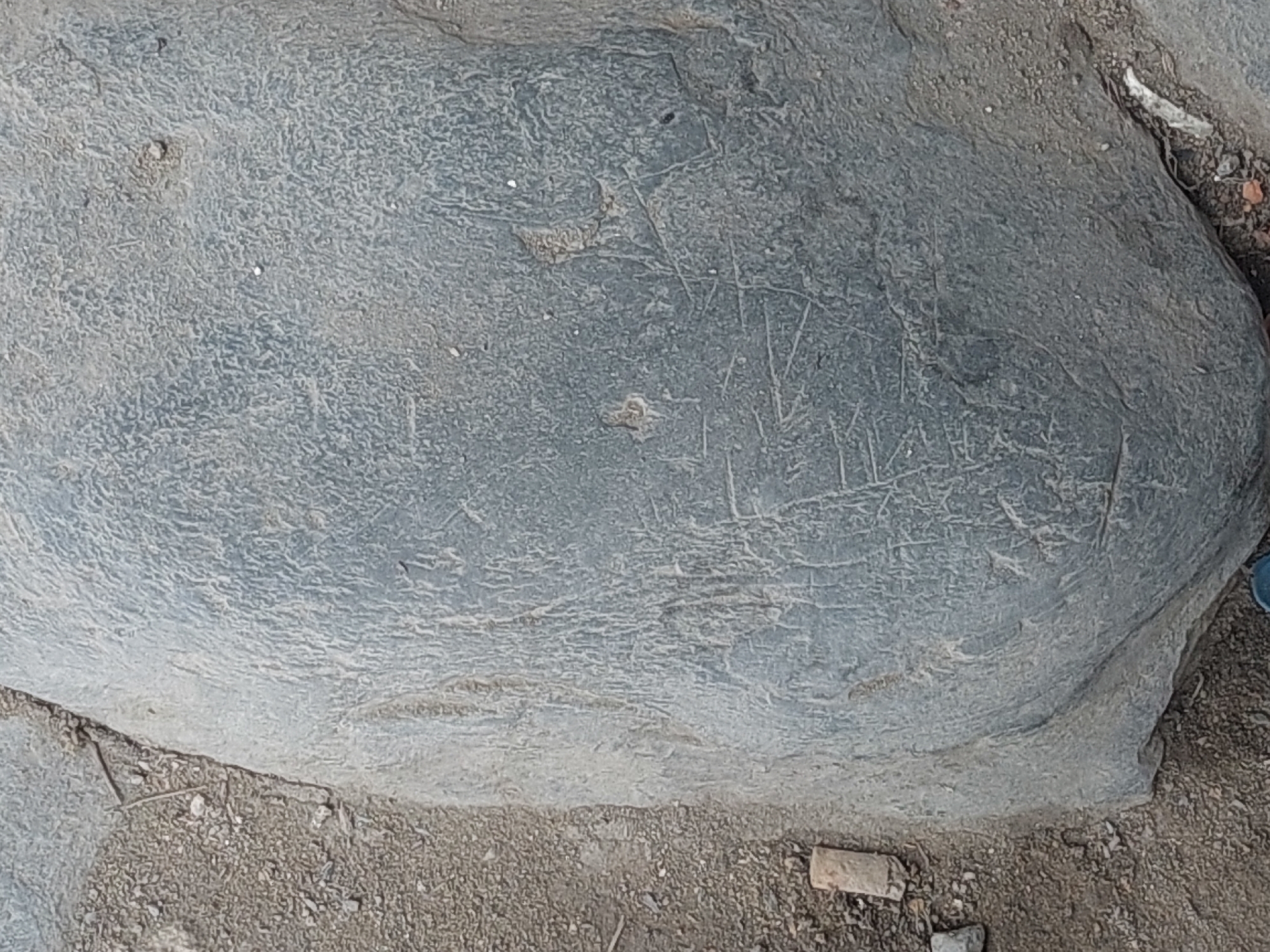 The image shows a large, gray, weathered rock with some faint markings etched into its surface. The rock is positioned on the ground, which is covered with dirt, small stones, and some debris. The environment appears to be outdoors, possibly in a natural or semi-developed area within the city of Parit Baru, Indonesia, given the context provided. There are no people visible in the image. The markings on the rock are indistinct, appearing as thin, scratched lines, possibly representing symbols or inscriptions, though their exact nature is not clear. The lighting suggests it is daytime, and the overall scene is one of an aged, static object in a simple outdoor setting.