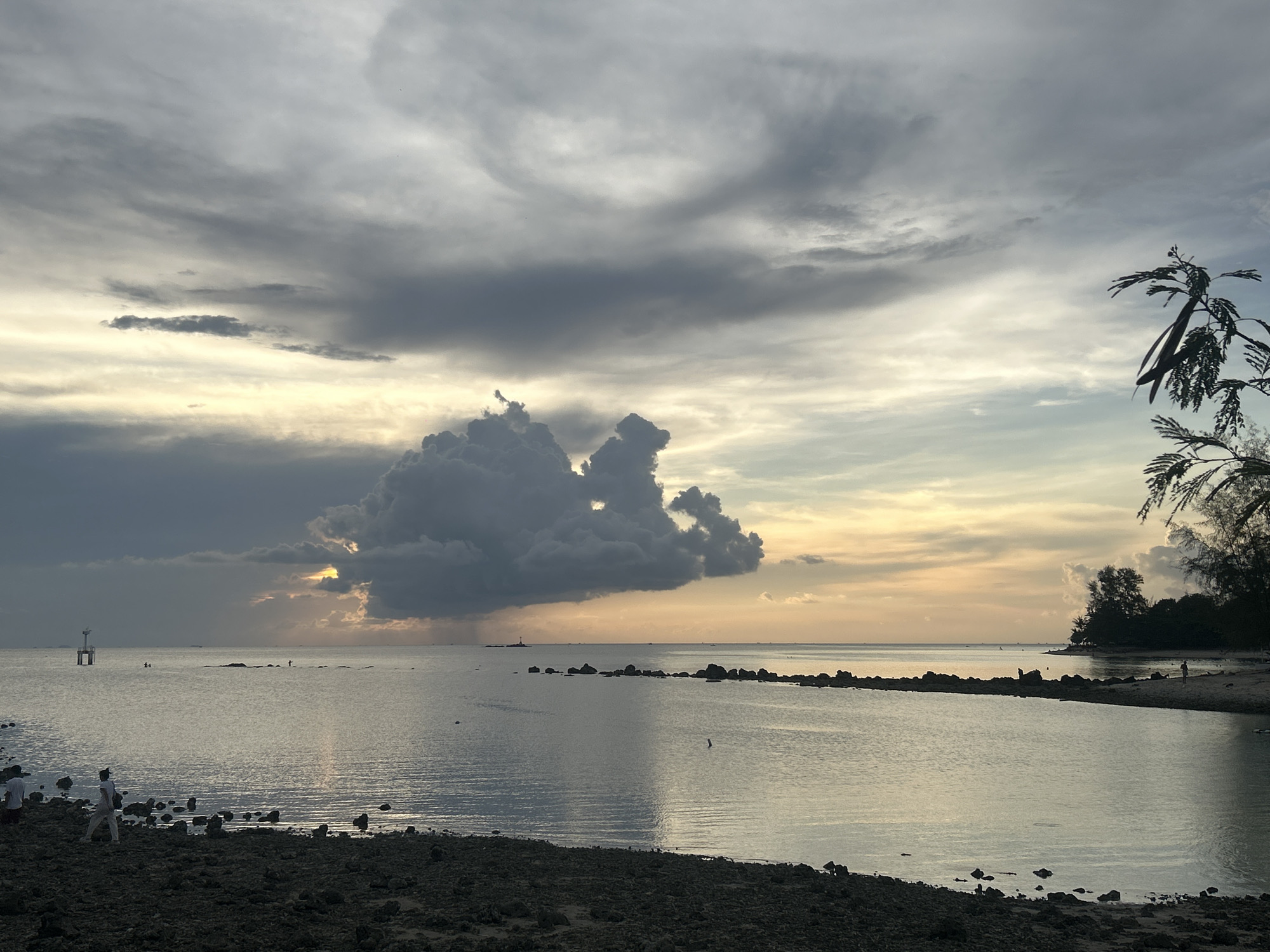 The image captures a twilight coastal scene in Baan Sri Thanu, Thailand. The sky is dominated by expansive cloud formations, ranging from swirling, high-altitude cirrus to a large, cumulonimbus mass. The sun is obscured, but light penetrates from behind the clouds, creating a diffused, golden glow on the horizon. The sea is calm, reflecting the sky and the sunlight. Along the shoreline, there is a rocky beach with scattered boulders and sparse vegetation. In the distance, there is a small island or headland covered with trees. A few people can be seen on the beach and in the water. There is a structure in the water to the left.
