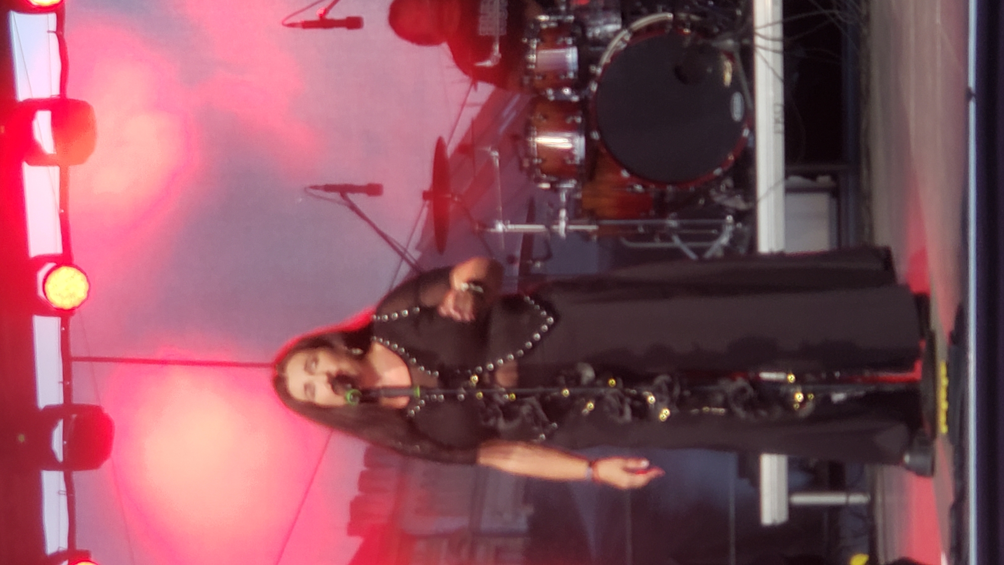A woman is singing into a microphone on a stage. She has long dark hair and is wearing a long black dress. She is lit by red stage lights. Behind her, a drum set and other stage equipment are visible. The image appears to be taken at an outdoor music festival in Prešov, Slovakia.
