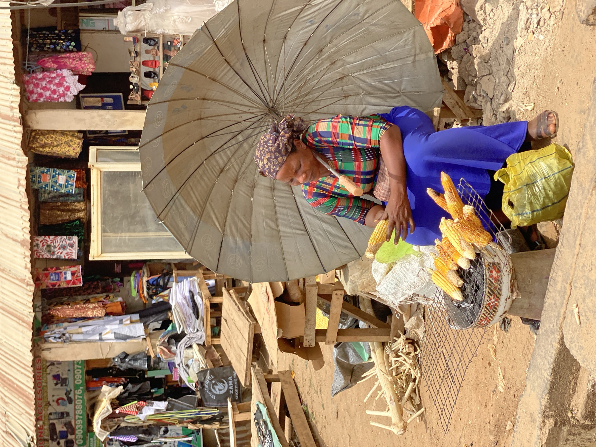This vertical shot captures a bustling outdoor market scene in what appears to be a sunny, daytime environment, likely in Kafanchan B, Nigeria. The central figure is a woman, possibly a vendor, seated on a low wooden structure under a large, light-colored umbrella that provides shade. She wears a colorful plaid shirt, a vibrant blue skirt or dress, and a patterned headscarf.
The woman is actively engaged in preparing or selling roasted corn. Several cobs of corn are visible on a makeshift grill, which consists of a metal grate placed over a container likely holding burning charcoal. She holds a knife with a wooden handle, perhaps used for shucking or serving the corn. Next to her, a bright yellow plastic bag rests on the ground. The immediate ground around her is rough, with dirt and some loose debris, including what looks like dried corn stalks or similar plant material.
In the background, behind and to the sides of the woman, are various market stalls and shops. These include displays of textiles, clothing, and footwear, suggesting a diverse marketplace. One sign is visible with text "PHONE ACCESSORIES" and phone numbers "09039788076" and "09073...". Another partially visible sign appears to have "NIG" on it. A "Wonderful Zama Shopping Bag" is also discernible in the background. The overall impression is that of an informal yet active commercial area, with goods displayed on wooden structures and shelves. The bright sunlight indicates good weather, and the umbrella is a practical necessity for the vendor.
