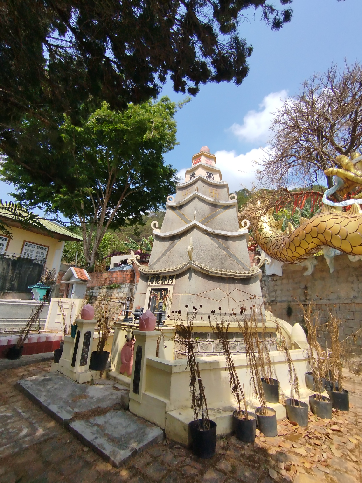 The image shows a traditional Vietnamese tomb located in what appears to be a cemetery. The tomb is a multi-tiered structure, predominantly off-white in color with gray accents, built in a style reminiscent of a pagoda. A gold-colored dragon statue is visible to the right of the tomb. A variety of trees are visible in the background. The image was taken in Diom A, Vietnam.