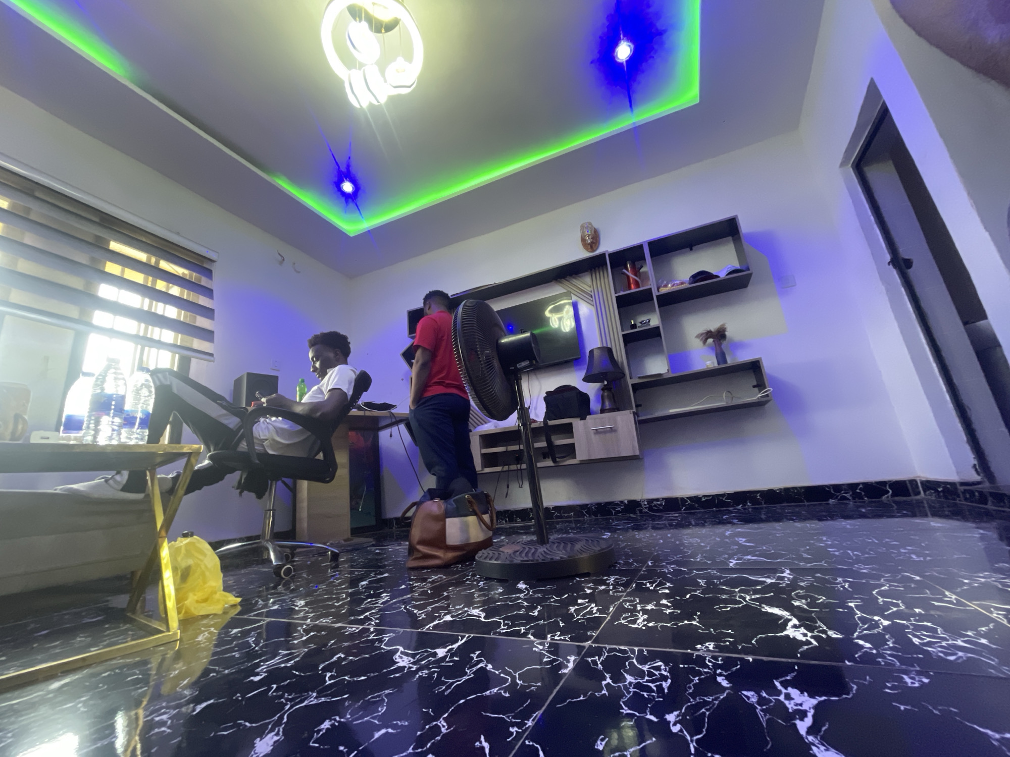This image depicts a modern indoor setting, likely a living room or studio, in Abuja, Nigeria, featuring two men. The room has a contemporary design with a striking black, highly polished floor exhibiting white marble-like veining, which reflects the overhead lights.
On the left, one man is seated in a black office chair at a desk, appearing to be focused on a laptop or screen. Several water bottles are visible on a nearby gold-framed side table, and a yellow plastic bag rests on the floor underneath it. A large window with horizontal blinds allows natural light to enter the room.
Towards the center-right, another man is standing near a tall, black standing fan, with his back partially to the camera. A multi-colored handbag is placed on the floor beside him. In the background, a flat-screen television is mounted above a sleek, dark wood wall unit with open shelves, displaying various decorative objects.
The ceiling features elaborate lighting: a large, modern white chandelier, green LED strip lights integrated into a ceiling cove, and blue spotlights that add a vibrant, colorful ambiance to the room. The overall impression is one of a stylish and functional space, with a mix of natural and artificial light sources creating a dynamic environment. No visible text is clearly legible in the image.