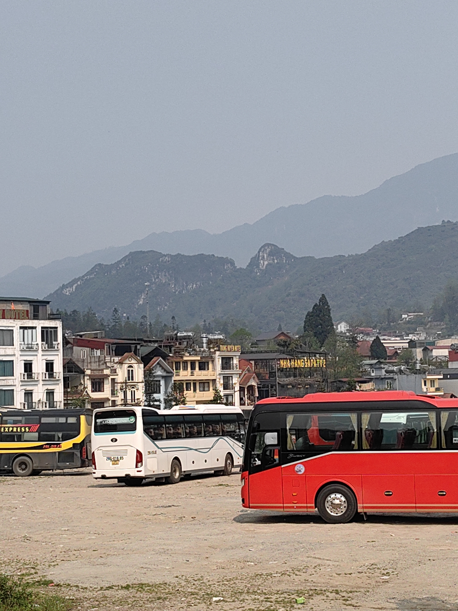 The image depicts a bus parking lot in Sa Pa, Vietnam, with a backdrop of urban structures and hazy mountains. The foreground is an unpaved, light brown dirt and gravel lot, with sparse green vegetation visible at the bottom left.
Three coach buses are parked in the lot, oriented towards the left. In the right foreground is a modern red coach bus, visible from its right side. It has black-tinted windows with light-colored curtains partially visible inside. The rear wheel features a silver rim. In the center midground, a white coach bus with blue/green accents is parked, displaying the Vietnamese license plate "29C-016.85" and the word "LIMOUSINE" on its rear. Reflections of passengers are faintly visible through its windows. To the left of the white bus, a black and yellow coach bus with "EXPRESS" and "PHÚ CA" text is partially visible from its right side. All buses appear stationary.
Behind the parking lot, a cluster of multi-story buildings forms the cityscape. On the far left, a white building with "HOTEL" marked in red features multiple balconies. Adjacent buildings vary in color (brown, yellow, white) and architectural style, with visible windows and various roof structures. A prominent yellow sign on a building in the center-right midground reads "NHA HANG SA PA TV2."
The background consists of several layers of green, tree-covered mountains, which fade into a light grey, hazy sky. A communication tower or antenna is discernible on one of the mountain slopes. No specific human actions or interactions are clearly depicted.