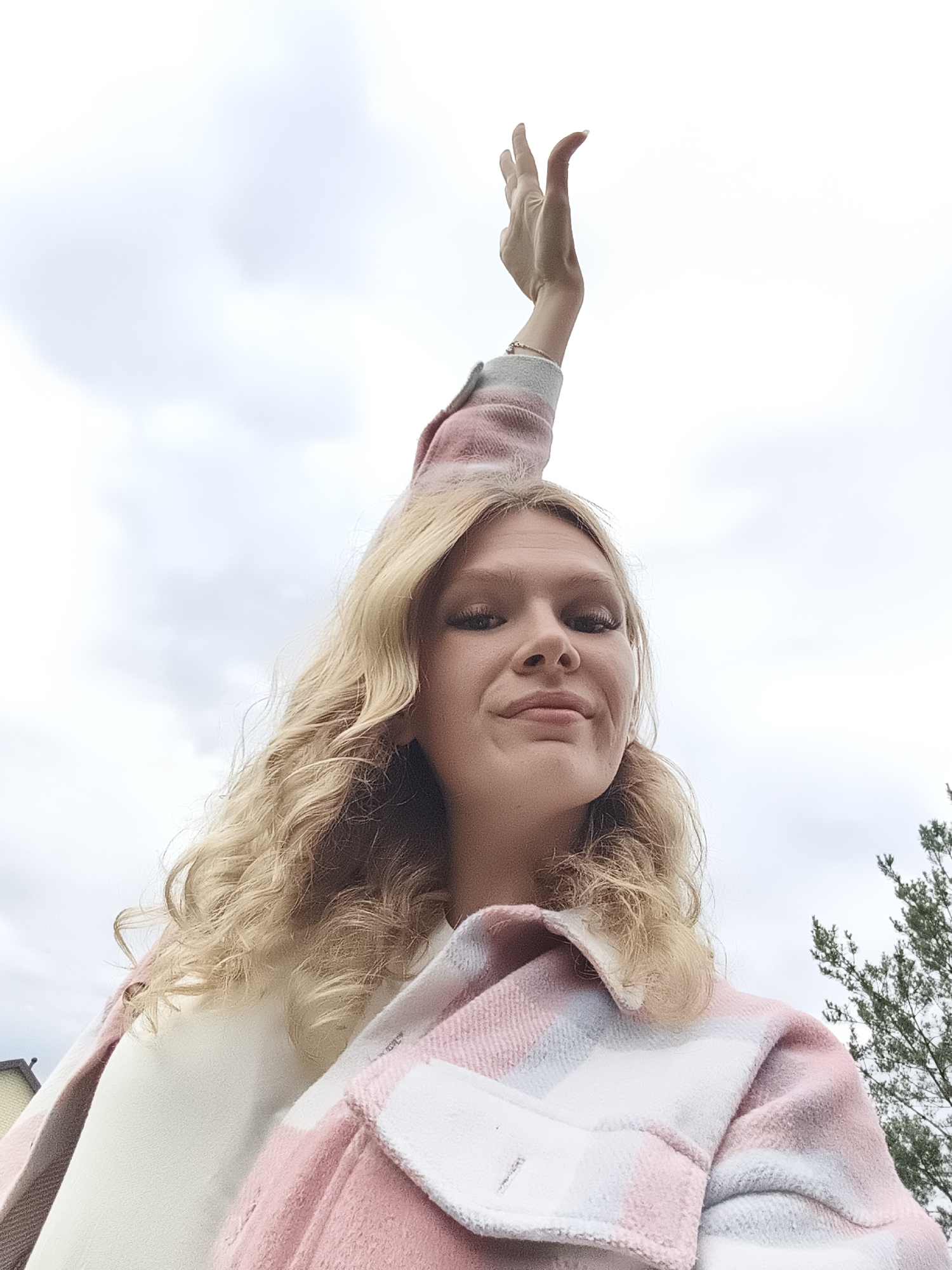 This is a low-angle, waist-up, outdoor selfie of a young woman with blonde, wavy hair. She is wearing a pink and white plaid shirt jacket over a cream-colored top. Her left arm is raised high, with her hand open and palm facing upwards. She is looking directly at the camera with a slight smile. The background is a cloudy sky, with some green foliage visible on the right side of the frame, suggesting a natural setting. The overall mood of the image is cheerful and dynamic. The time of day appears to be daytime, given the brightness of the sky, and the weather is overcast. There is no visible text in the image.