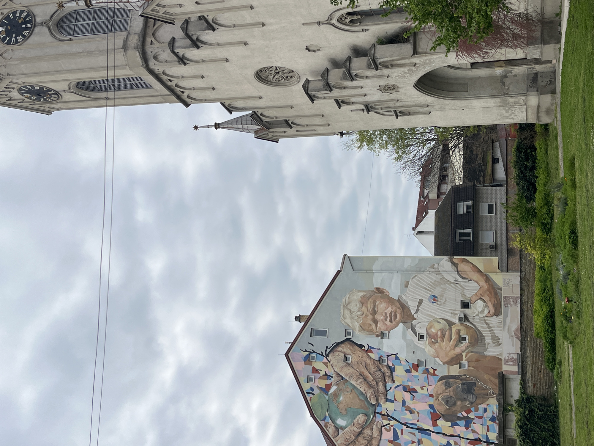 The image shows a view in Novi Sad, Serbia, featuring a building with a large mural juxtaposed with the iconic tower of the Name of Mary Church (Crkva Imena Marijinog). The mural depicts two elderly figures and a dog; the lower figure’s hands are holding a globe. The mural is on the side of an apartment building. The sky is cloudy. Other buildings and greenery are visible in the background.