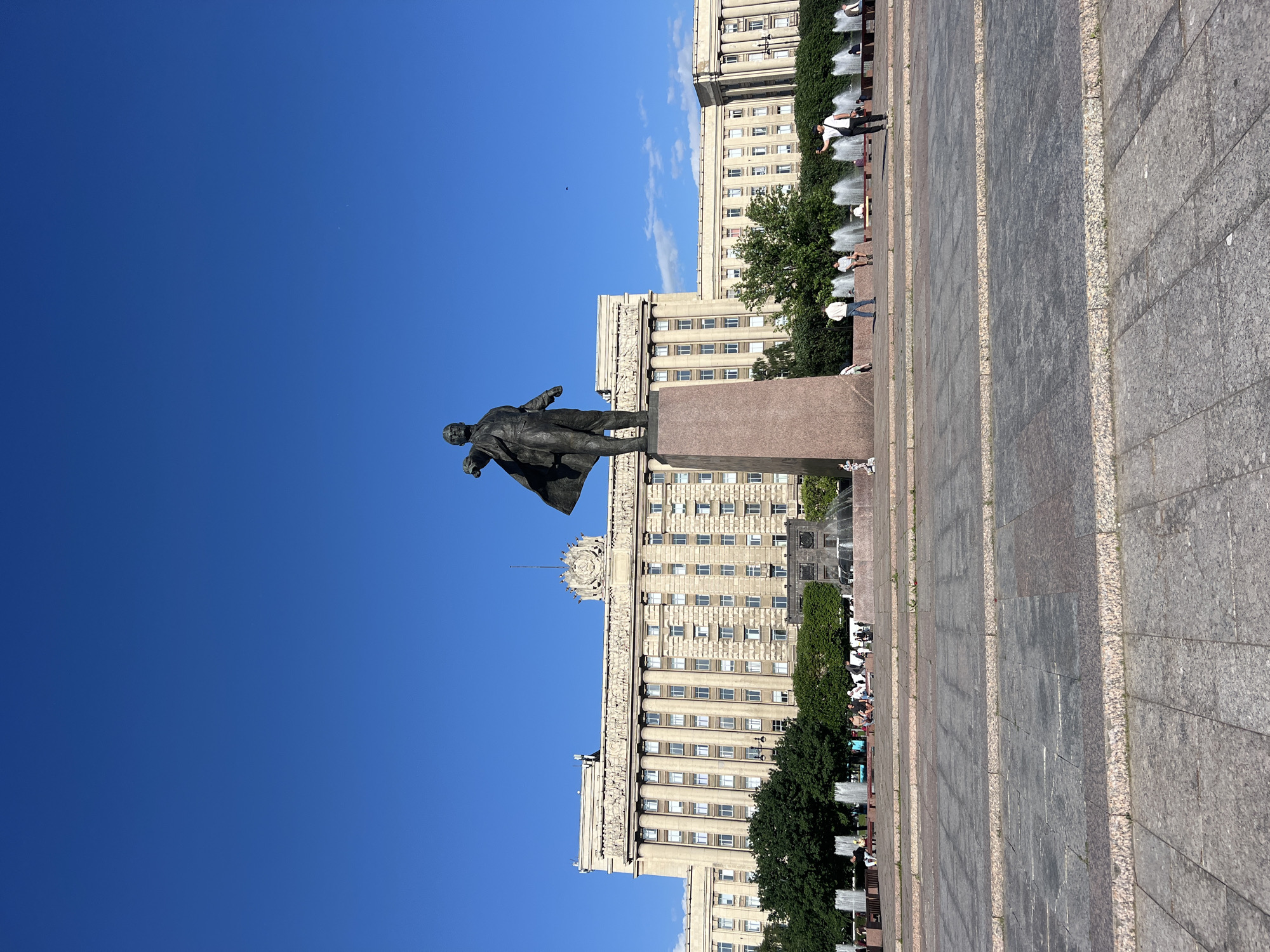 The image depicts a wide, paved plaza in front of a large, classical-style building with columns and many windows. A tall bronze statue of Vladimir Lenin, a prominent figure in Russian history, stands on a pedestal in the foreground, facing to the left and gesturing with his outstretched arm. Behind Lenin, a row of fountains sprays water into the air. Several people are visible in the plaza, some sitting on benches, others walking, and one person is taking a selfie. Lush green trees line the background, providing a contrast to the stone architecture and paving. The sky above is a clear, vibrant blue. This scene is of the Lenin monument in front of the Smolny Institute in Saint Petersburg, Russia.