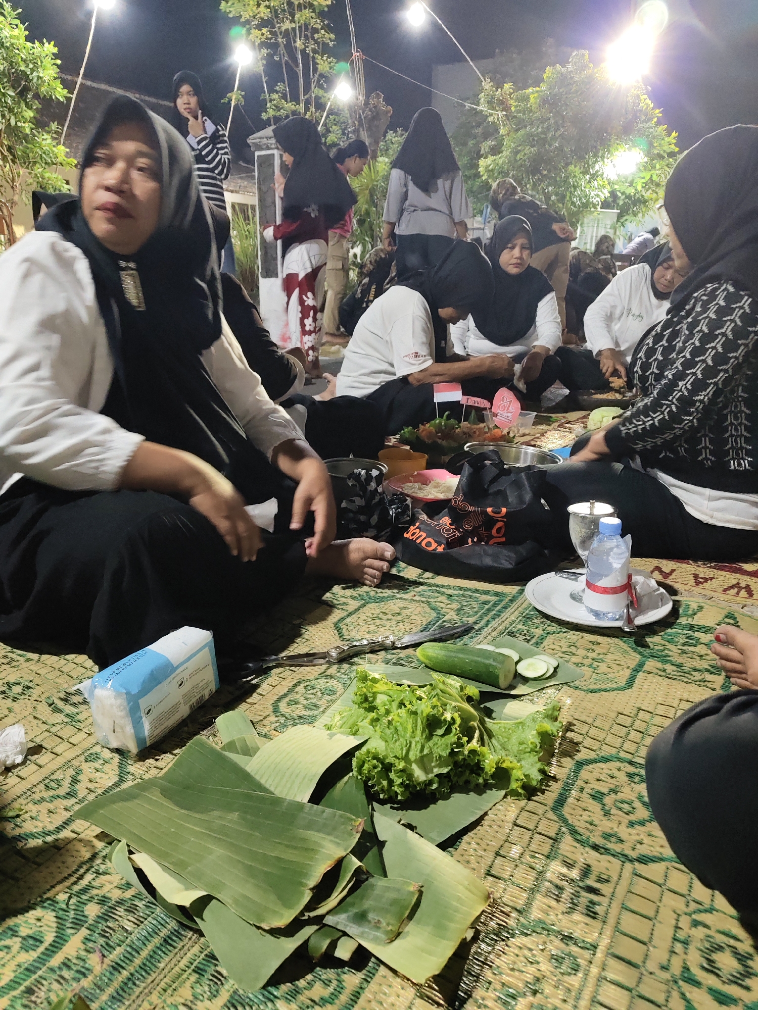 In Gumpang, Indonesia, a group of people are gathered outdoors at night, likely for a meal or a social event. The scene is illuminated by overhead lights, casting a warm glow on the participants. Most individuals are wearing hijabs, indicating a cultural context where this is customary. Some are seated on a woven mat, while others stand or are partially visible in the background.
In the foreground, a woman in a black hijab and white top is seated, her gaze directed away from the camera. To her right, another person, also in a dark hijab and white top, is engaged with something on the mat. The mat is covered with food items, including fresh lettuce, sliced cucumber on banana leaves, and what appears to be a packet of tissues and a bottle of water. A knife and scissors are also visible on the mat. In the middle ground, a black bag with orange lettering and other food containers are present. Several small flags, including what looks like an Indonesian flag, are stuck into a dish of food.
The background shows other individuals, some with hijabs, and trees with illuminated foliage. The overall atmosphere suggests a relaxed, communal gathering.