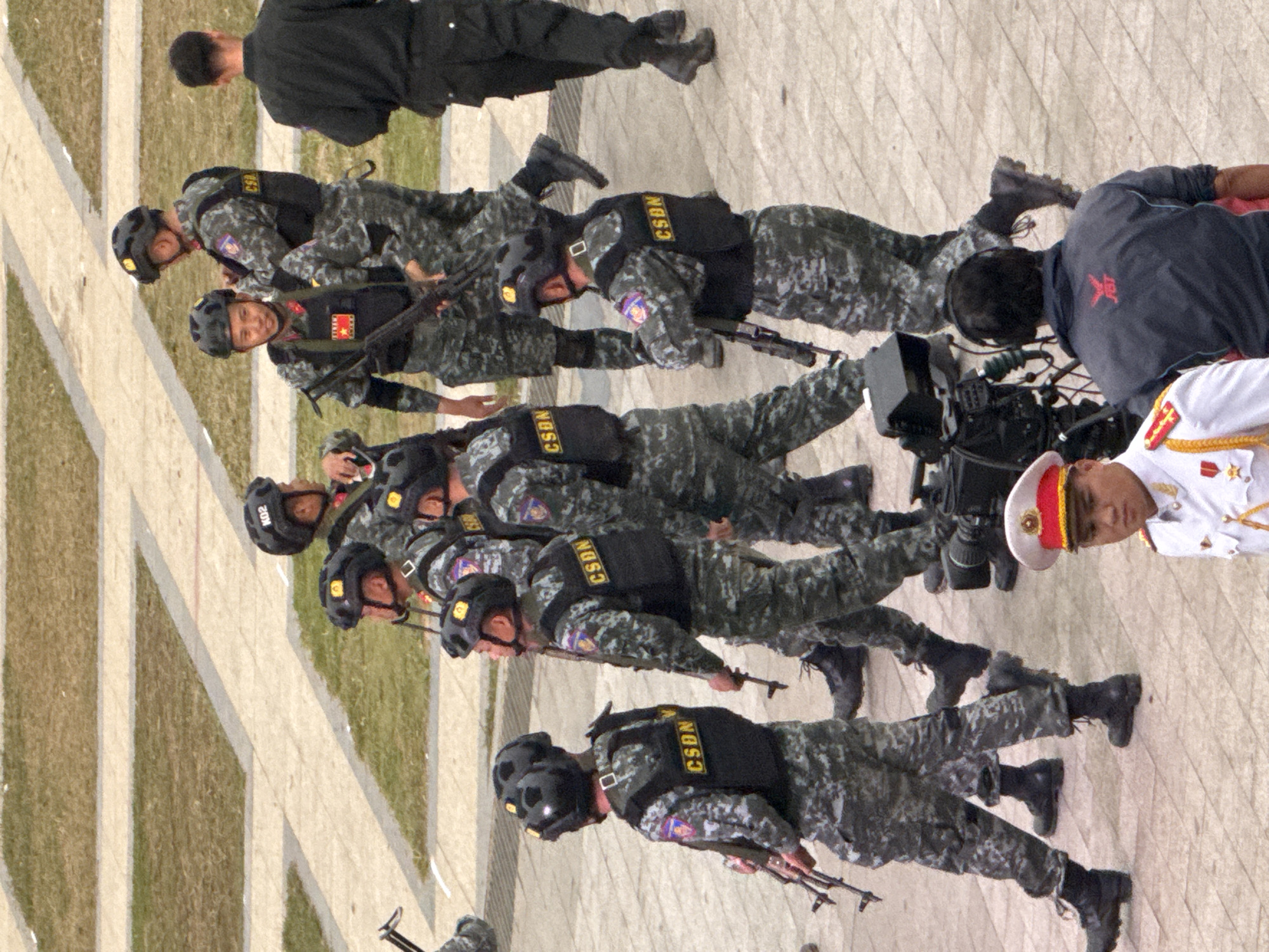 The image shows a group of people in military uniforms, standing in formation. The subjects are mostly men, dressed in camouflage uniforms, tactical vests, and helmets. Some are holding rifles. In the foreground, a person in a white uniform with a red and gold hat is also visible. The setting appears to be outdoors, with a paved ground and some grassy areas. The people are arranged in a diagonal line, suggesting a parade, ceremony, or training exercise. The lighting suggests it is daytime. The orientation of the image is rotated, with the people appearing to be standing upright when viewed in the correct orientation. Visible text on the vests includes "CSDN" and what appears to be a national emblem. The location context provided is Da Lat, Vietnam, which might imply the event is related to a Vietnamese military or police event. The expressions on the faces of the individuals are neutral, consistent with a formal military posture.
