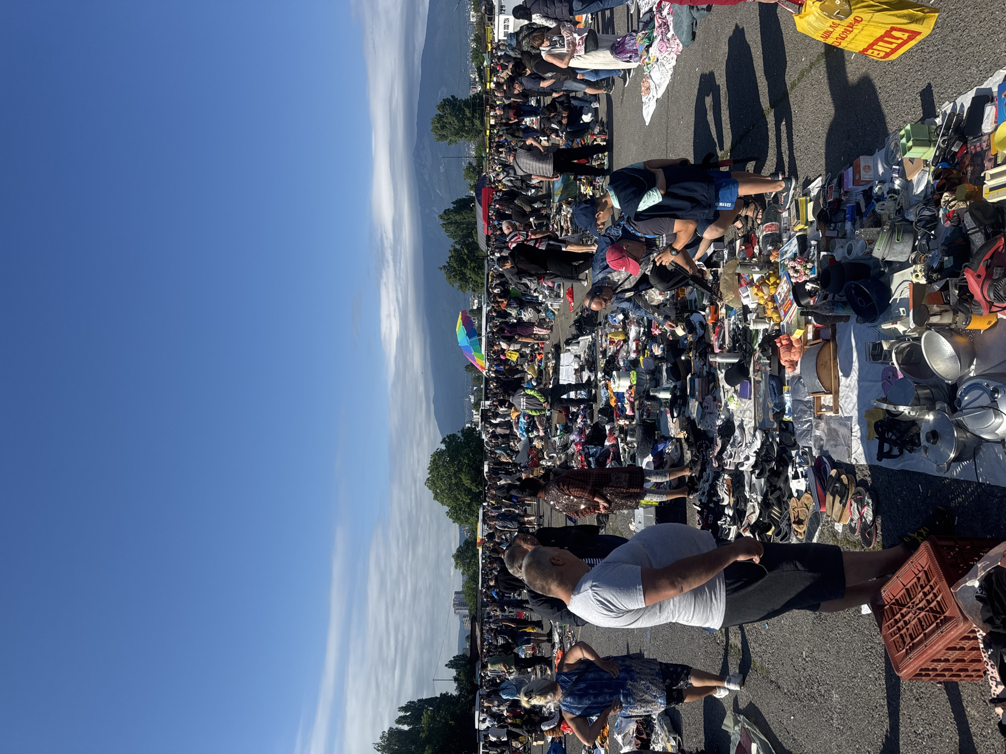 This is a wide shot of an outdoor market, likely a flea market or car boot sale, taking place on a paved surface under a clear blue sky with distant mountains. The market is crowded with people browsing and shopping at numerous stalls laden with a variety of goods, including clothing, shoes, kitchenware, tools, and miscellaneous items. The general atmosphere appears busy and lively. In the background, there are trees, buildings, and the distinctive silhouette of Vitosha Mountain, a prominent landmark of Sofia, Bulgaria. A colorful umbrella is visible in the distance, adding a splash of color to the scene. The lighting suggests it is a sunny day, likely late morning or early afternoon.