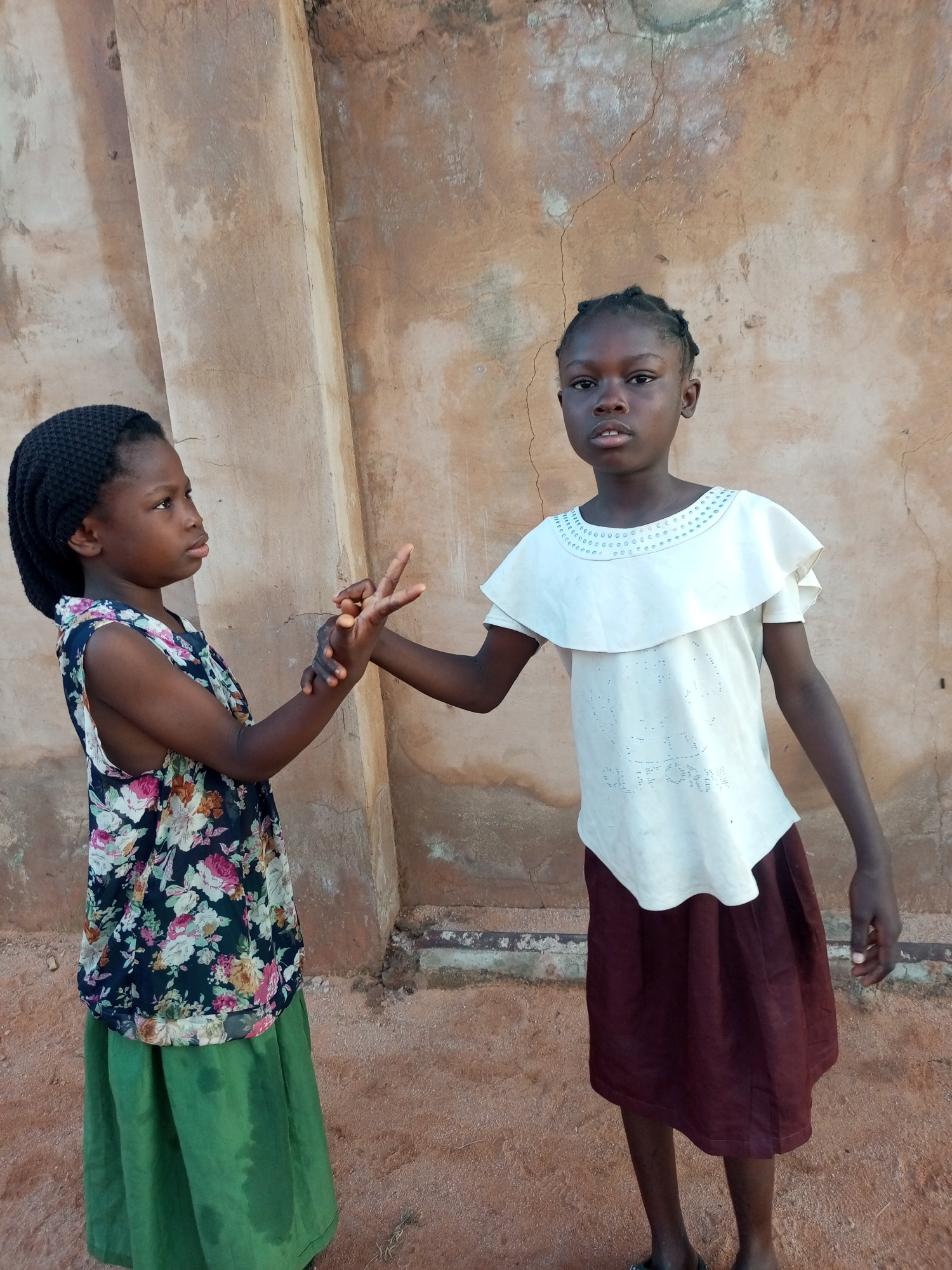 Two young girls stand in front of a textured, tan-colored wall in Tempure, Nigeria. The girl on the left wears a black headscarf and a dark floral-patterned sleeveless top over a long green skirt. She is looking to her right and has her left hand reaching out towards the other girl. The girl on the right wears a white ruffled top with light blue embellishments around the neckline and a dark red skirt. Her right hand is raised, and her fingers are splayed, with her index and middle fingers extended in a peace sign gesture, touching the left girl's outstretched hand. The ground beneath them is sandy and reddish-brown. The wall behind them is rough and appears weathered, with some vertical lines or cracks.