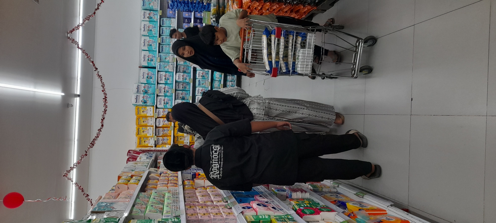 In a brightly lit supermarket aisle, possibly located in Situbondo, Indonesia, a group of people are seen browsing. A young boy, wearing a light green t-shirt, pushes a shopping cart filled with packaged goods, some wrapped in blue and white. Beside him, a woman in a black hijab and a patterned dress observes the products. Behind them, a man in a black t-shirt with a logo that reads "Rognocs" stands with his back to the camera, wearing dark pants and sandals. Another person in a black hijab is partially visible behind the man. The shelves on either side of the aisle are stocked with various consumer products, predominantly diapers and other packaged items. A red balloon is suspended from the ceiling, attached to a string that trails downwards, adding a festive touch. Decorative red and white garlands are strung along the ceiling. The floor is tiled with light-colored squares.