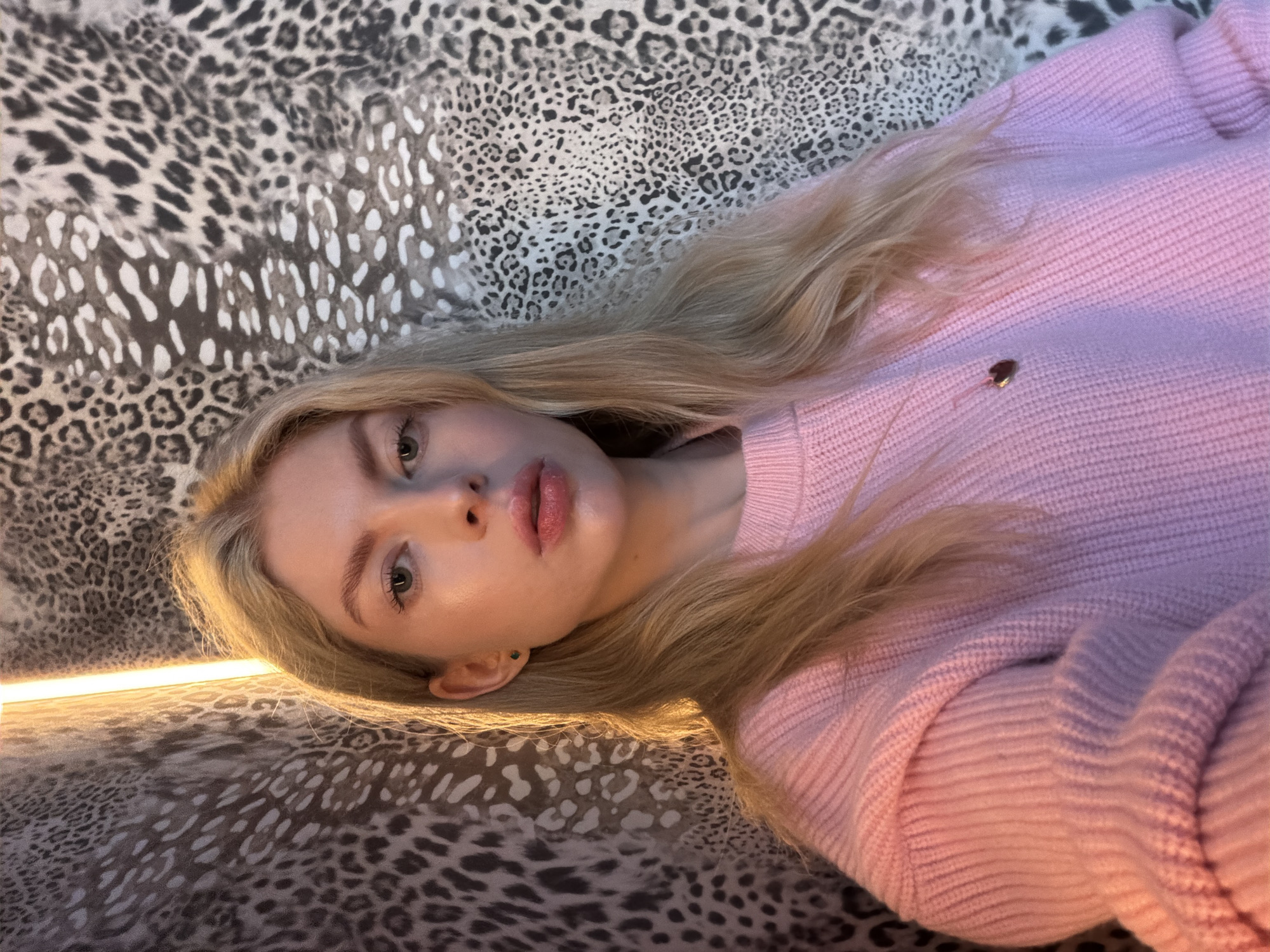 A young woman with blonde hair and light skin is taking a selfie. She is wearing a pink knitted sweater and a heart-shaped pendant necklace. The background is a wall with a leopard print pattern, and a vertical light source is illuminating the scene from the left. The woman is positioned in the center of the frame, with her head tilted slightly to her left. Her eyes are open and looking towards the camera. Her lips are slightly parted. The lighting suggests an indoor setting, possibly a home or a studio. The image does not contain any specific geographical indicators that would allow for identification of a location within Moscow, Russia.