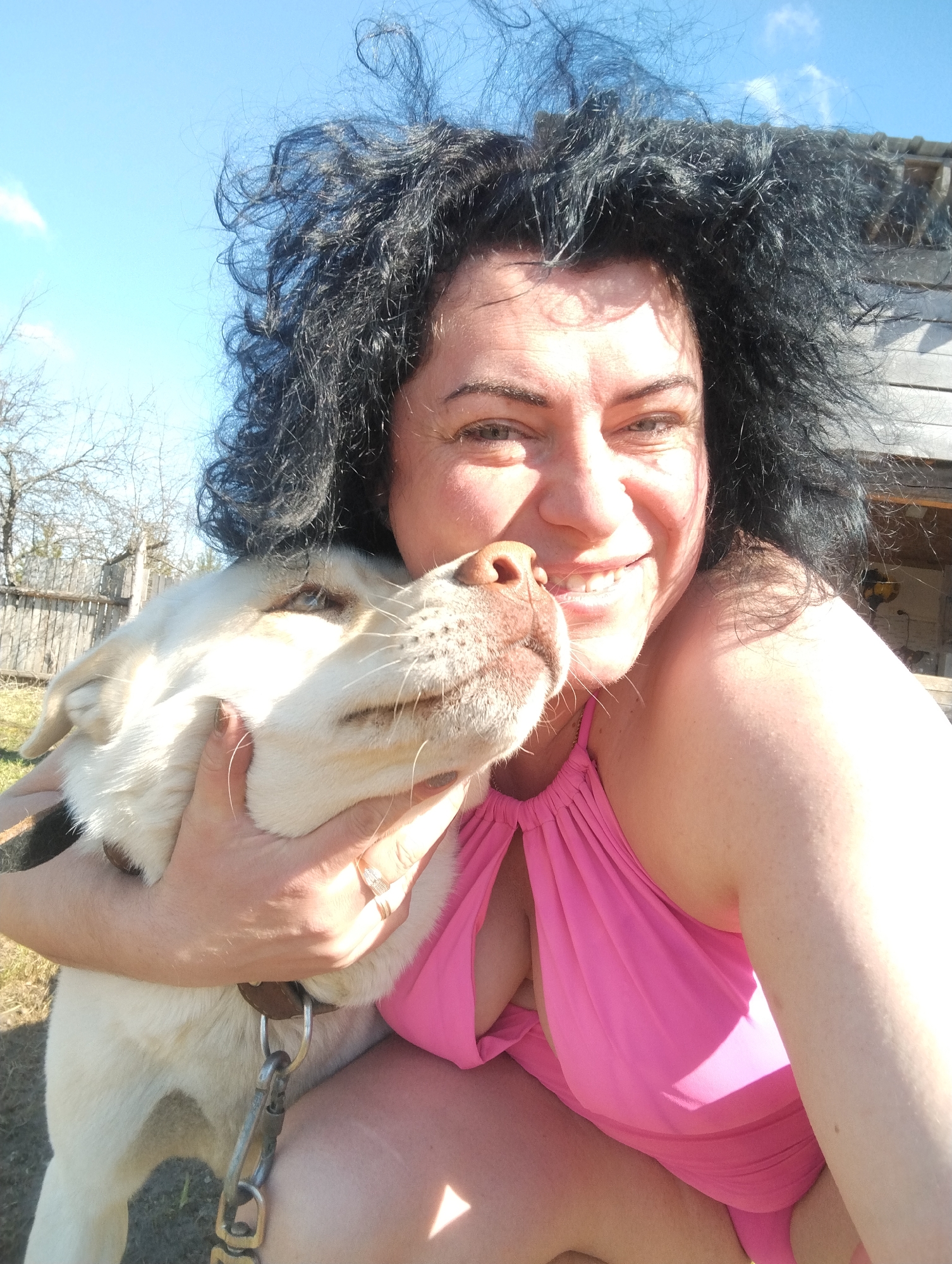 An adult woman and a light-colored dog are captured in a close, affectionate embrace outdoors in Grishanki, Russia. The woman, with voluminous dark curly hair, is smiling broadly, her face lit by bright sunlight. She is wearing a vibrant pink halter-neck top or swimsuit. Her left arm is wrapped around the dog, holding it close to her. Visible rings adorn her left hand.
The dog, which appears to be a large breed like a Labrador with light cream or white fur, has its head resting against the woman, looking up at her with a gentle expression. It wears a brown leather collar with a metal leash clip attached.
The activity depicts a moment of strong bond and affection between the woman and her pet. The time of day is clearly daytime, indicated by the bright, direct sunlight and visible shadows. The weather is sunny with a clear blue sky and a few wispy clouds. The setting appears to be a rural or semi-rural outdoor area, with bare tree branches, a wooden fence, and part of a rustic wooden building visible in the background. The ground is a mix of grass and dirt. Both subjects convey emotions of happiness and contentment. No visible text is present in the image.