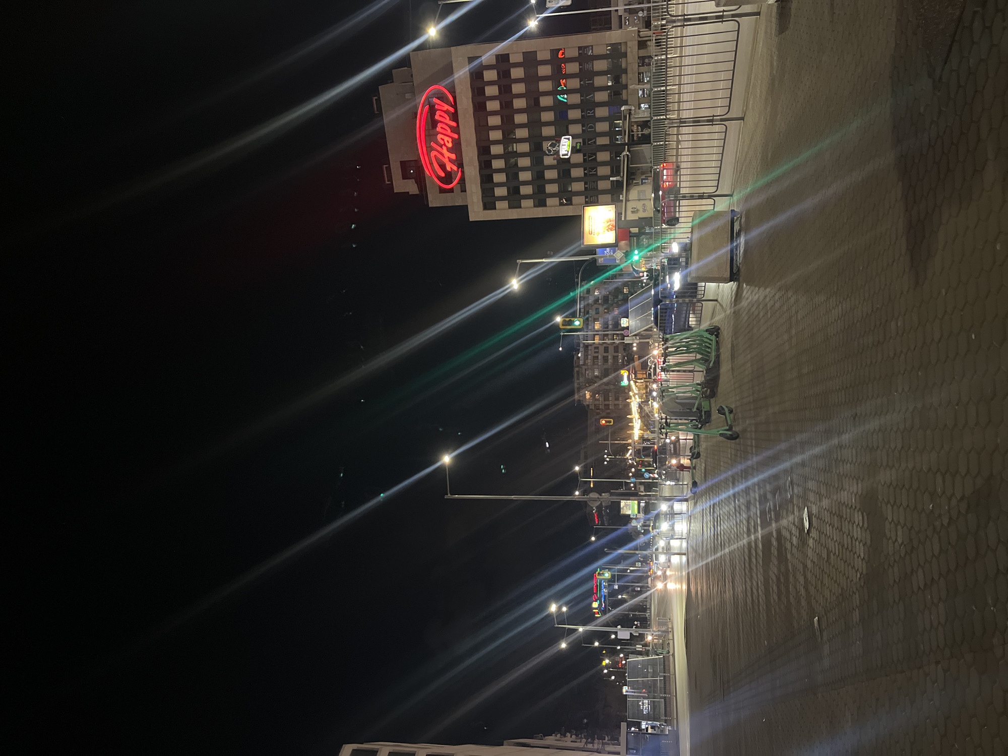The image depicts a night scene on a street in Sofia, Bulgaria. The distinct red "Happy" restaurant sign is visible on a building. Streetlights illuminate the scene, creating bright flares. A row of scooters is parked on the side of the street. Cars are visible in the distance. The ground is paved with hexagonally patterned bricks. A metal railing runs along the sidewalk.