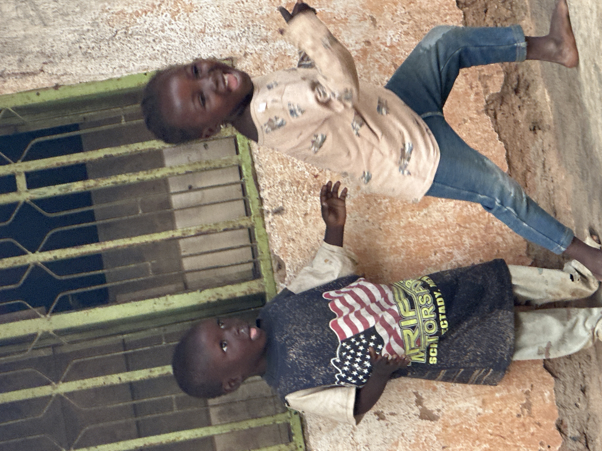 Two young children, likely of African descent, stand in front of a textured, orange-brown wall. The child on the left, wearing a t-shirt with the American flag graphic and a dark blue overlay, has their tongue sticking out. The child on the right, in a light-colored long-sleeved shirt and blue jeans, is laughing with their mouth open wide and pointing their right index finger forward. A green-barred window is visible behind them. The ground appears to be dirt or concrete. The scene is likely located in Gura Top, Nigeria.