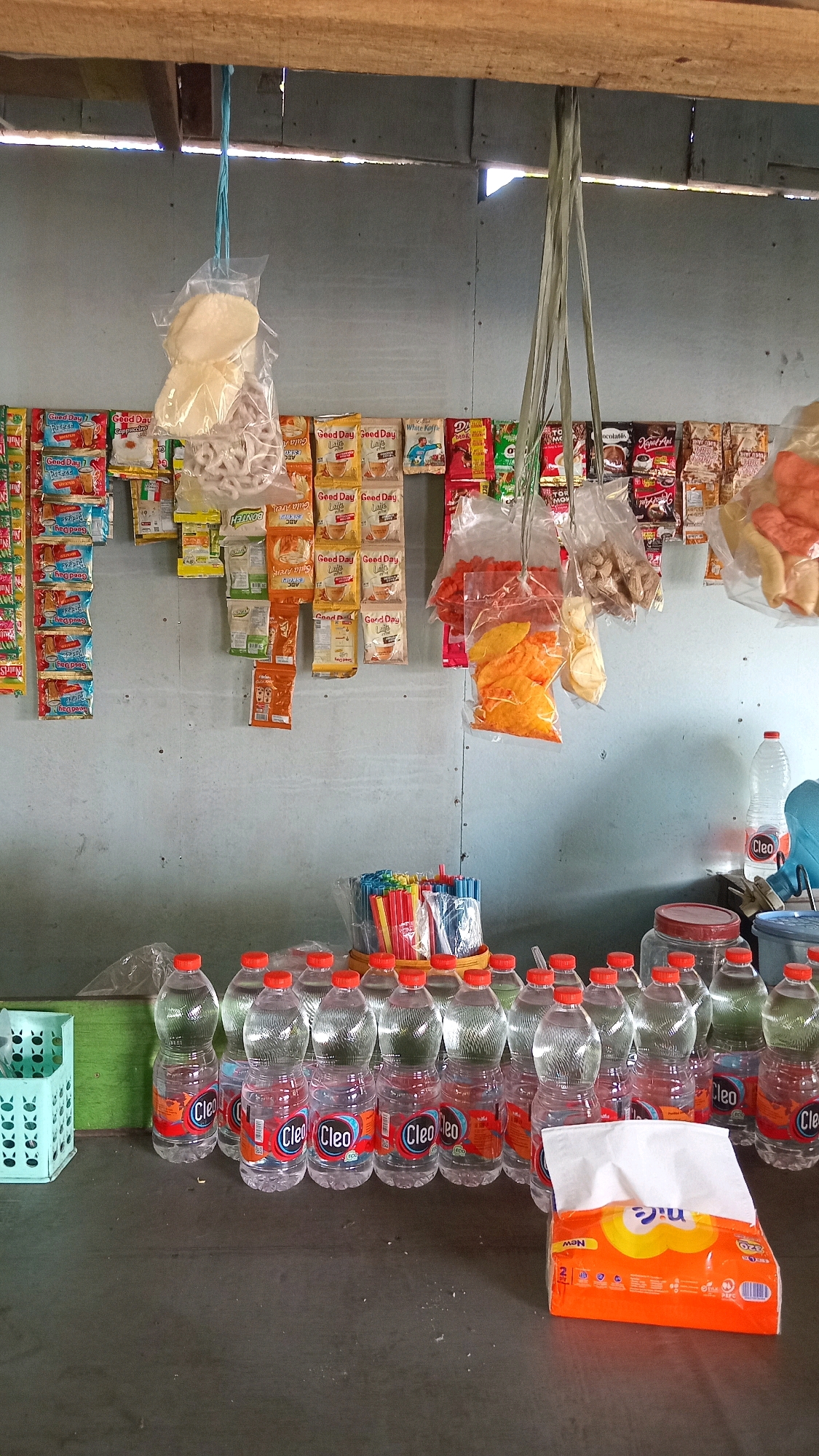 This image depicts a small roadside stall in Ambeng Ambeng Watangrejo, Indonesia. The stall features a variety of packaged goods hanging from the ceiling and displayed on shelves and a counter. Items include snacks, instant coffee packets (such as "Good Day" brand), and bottled water. Various bottled drinks with orange caps are lined up on the counter. There are also plastic bags containing snacks, a box of tissues, and a container of straws. The stall has a rustic appearance, with a wooden beam supporting the roof and a corrugated metal wall behind the merchandise. The overall impression is that of a local convenience store or warung.