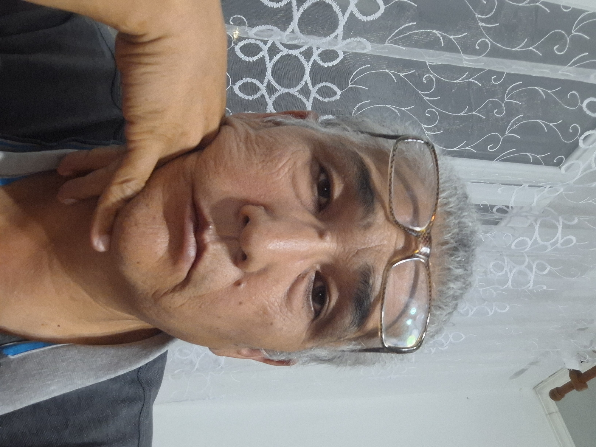 A close-up, head-and-shoulders selfie of an elderly East Asian man with graying hair and glasses is taken indoors. The man is resting his chin on his hand and looking directly at the camera. His face shows signs of age, with wrinkles around his eyes and mouth. He is wearing a dark gray shirt with a lighter gray collar. In the background, a sheer white curtain with a delicate floral pattern is visible. The overall lighting suggests an indoor setting, likely a home. There is no visible indication of the city of Almaty, Kazakhstan.