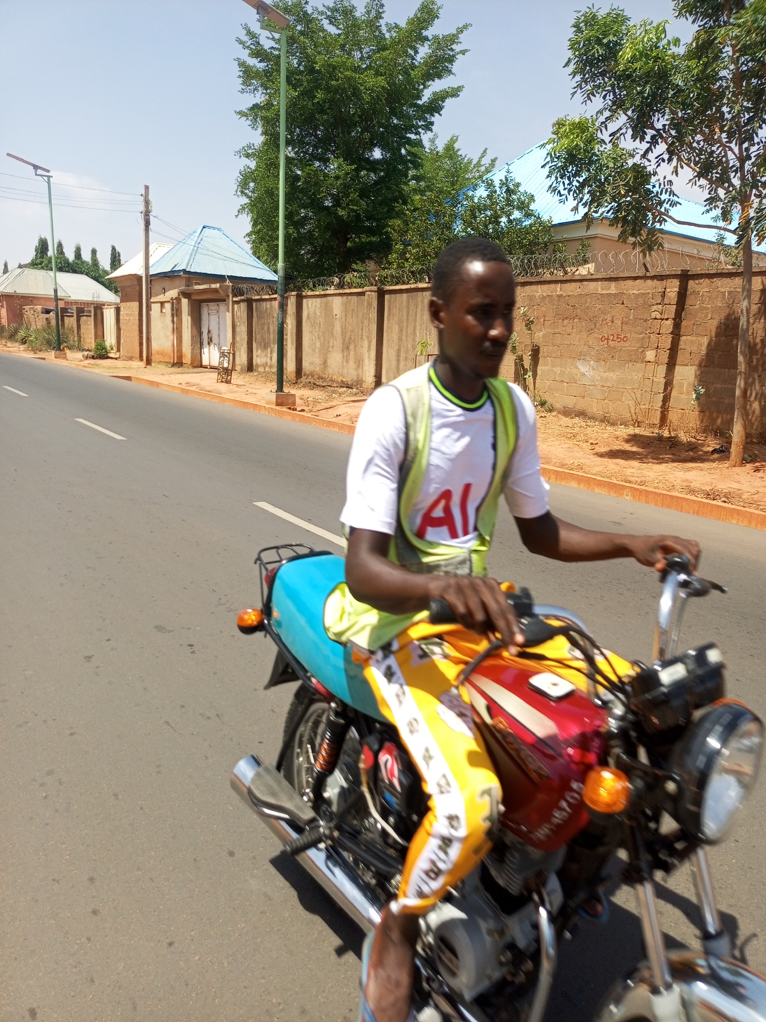 A man is riding a motorcycle on a paved road. The man is wearing a white T-shirt with a green collar, a yellow safety vest, and yellow pants with white stripes. The motorcycle has a blue seat and a red gas tank. The road has white lines and is bordered by a low wall. Behind the wall are buildings with blue roofs and trees. A tall street light is located on the side of the road. The location is Unknown, Unknown.