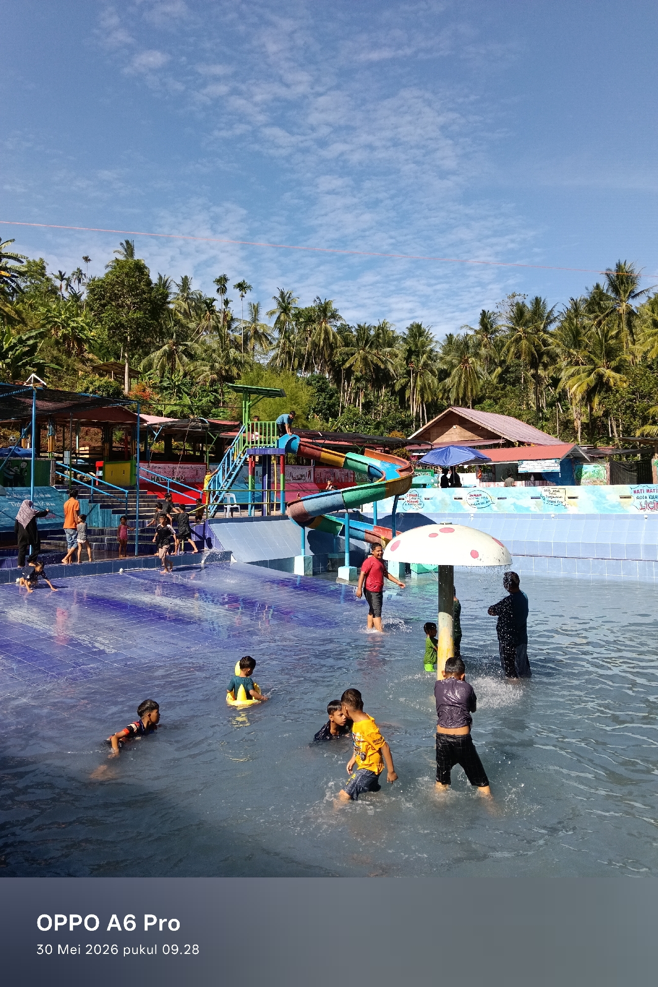 This photograph captures a scene at a water park, located in the city of Palohseulimeng, Indonesia. The park features a blue-tiled pool filled with people, mostly children, enjoying the water. Some are swimming, while others are standing and playing near a mushroom-shaped sprinkler. In the background, there's a colorful water slide structure with stairs leading up to the top. Surrounding the water park is lush greenery with tall trees, including coconut palms, adding to the tropical atmosphere. The sky above is bright blue with scattered white clouds. There are buildings and shelters, providing shade, along the perimeter of the pool. The text on the lower portion of the image reads "OPPO A6 Pro 30 Mei 2026 pukul 09.28". There is also a sign that has partially visible text, "HATI HATI AREA KOLA LICIL".