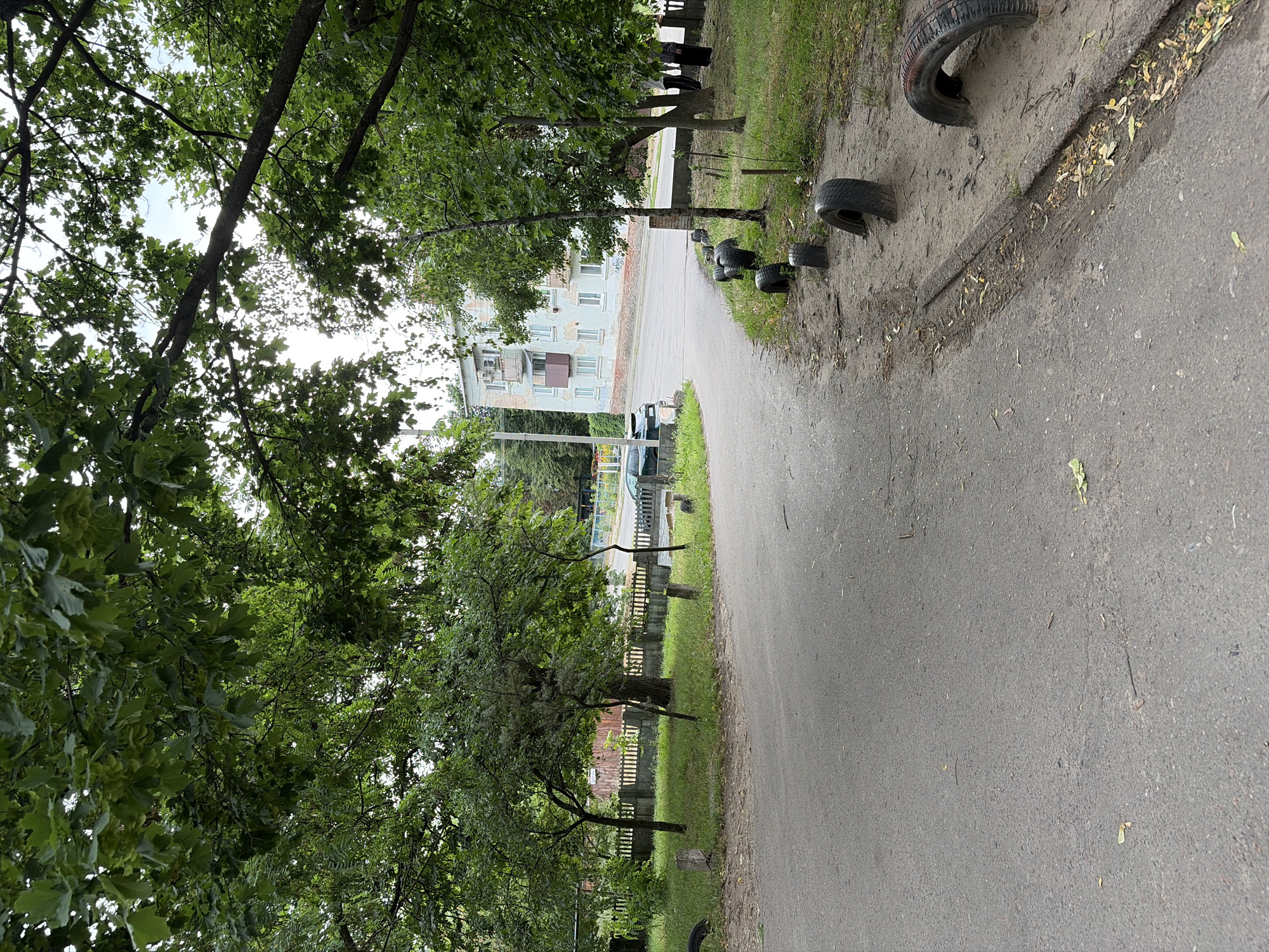 This media file captures an outdoor scene on a cloudy day, likely in Chernihiv, Ukraine. The image is vertically oriented and shows a pathway or road bordered by a grassy area and trees on the left. On the right side of the pathway, several old, worn-out tires are placed in a line along the edge of the sandy or dirt area. Beyond the pathway, a multi-story apartment building with light blue walls and brown balconies is visible, suggesting a residential area. The building appears to be of Soviet-era architecture.
The trees are lush and green, indicating it's likely spring or summer. The sky is overcast, and there's no direct sunlight, contributing to a slightly muted color palette. The pathway itself is paved and appears to be a road or a wide pedestrian path. There's a general sense of an urban or suburban environment, with elements of nature and weathered man-made structures.
No people are visible in the image, and no specific activity or event is depicted. The focus is on the environment, with the discarded tires as a notable detail that might suggest a playground area, a makeshift barrier, or simply refuse. The overall mood is neutral, with no strong emotional cues conveyed. The presence of the apartment building and the paved pathway points towards a community setting.