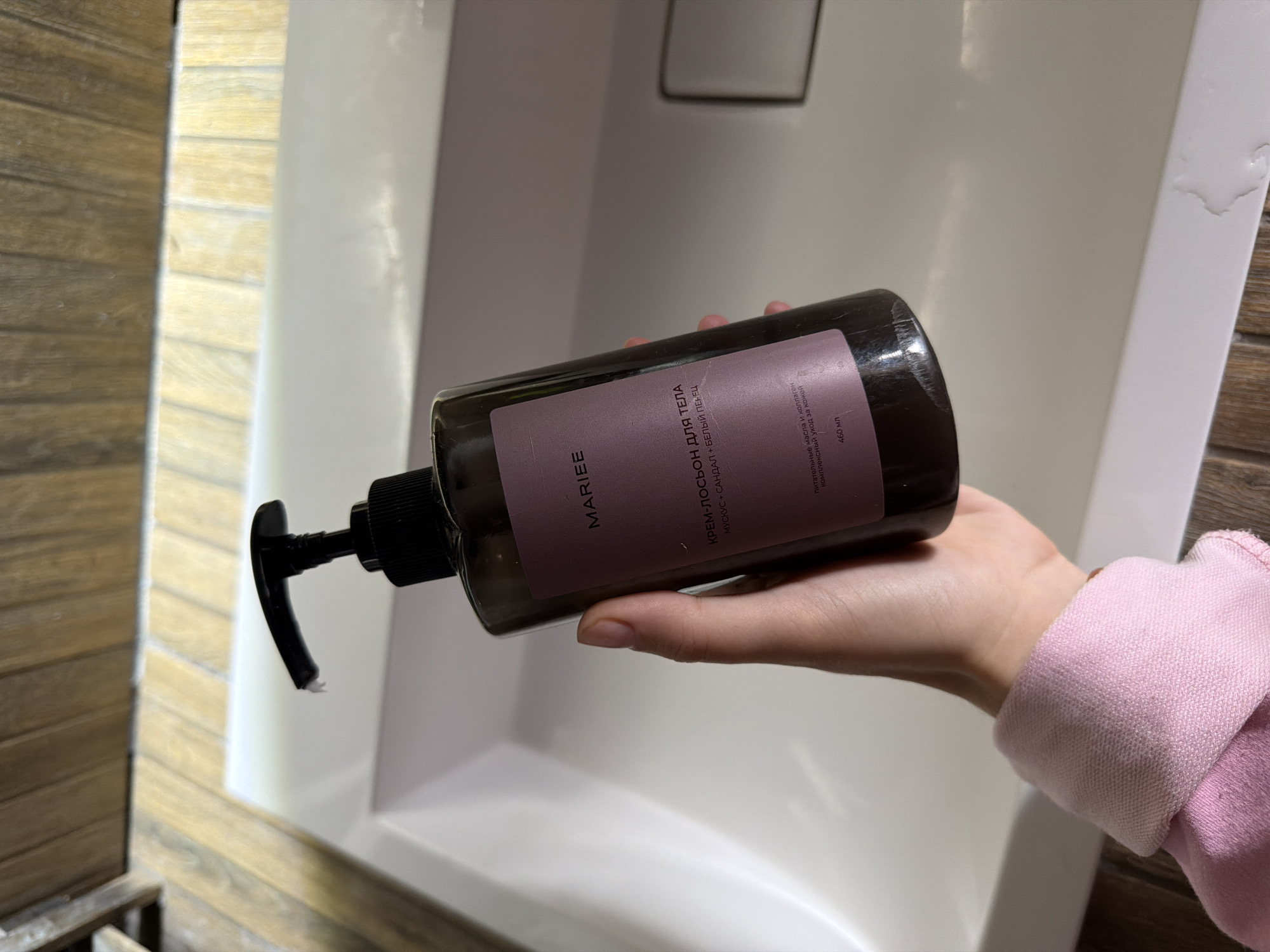 A hand with light skin and short fingernails holds a dark brown, translucent bottle of "MARIEE" body cream-lotion with a pump dispenser. The bottle's label is dark purple and has white text. The text on the label reads: "MARIEE", "КРЕМ-ЛОСЬОН ДЛЯ ТЕЛА", "МУСКУС + САНДАЛ + БЕЛЫЙ ПЕНЕЦ", and "ПИТАТЕЛЬНЫЕ МАСЛА И КОЛЛАГЕН КОМПЛЕКСНЫЙ УХОД ЗА КОЖЕЙ", and "460 мл". The background is a white sink with wooden accents above it. This scene does not provide enough information to determine its location in the city of Семилуки, Russia.