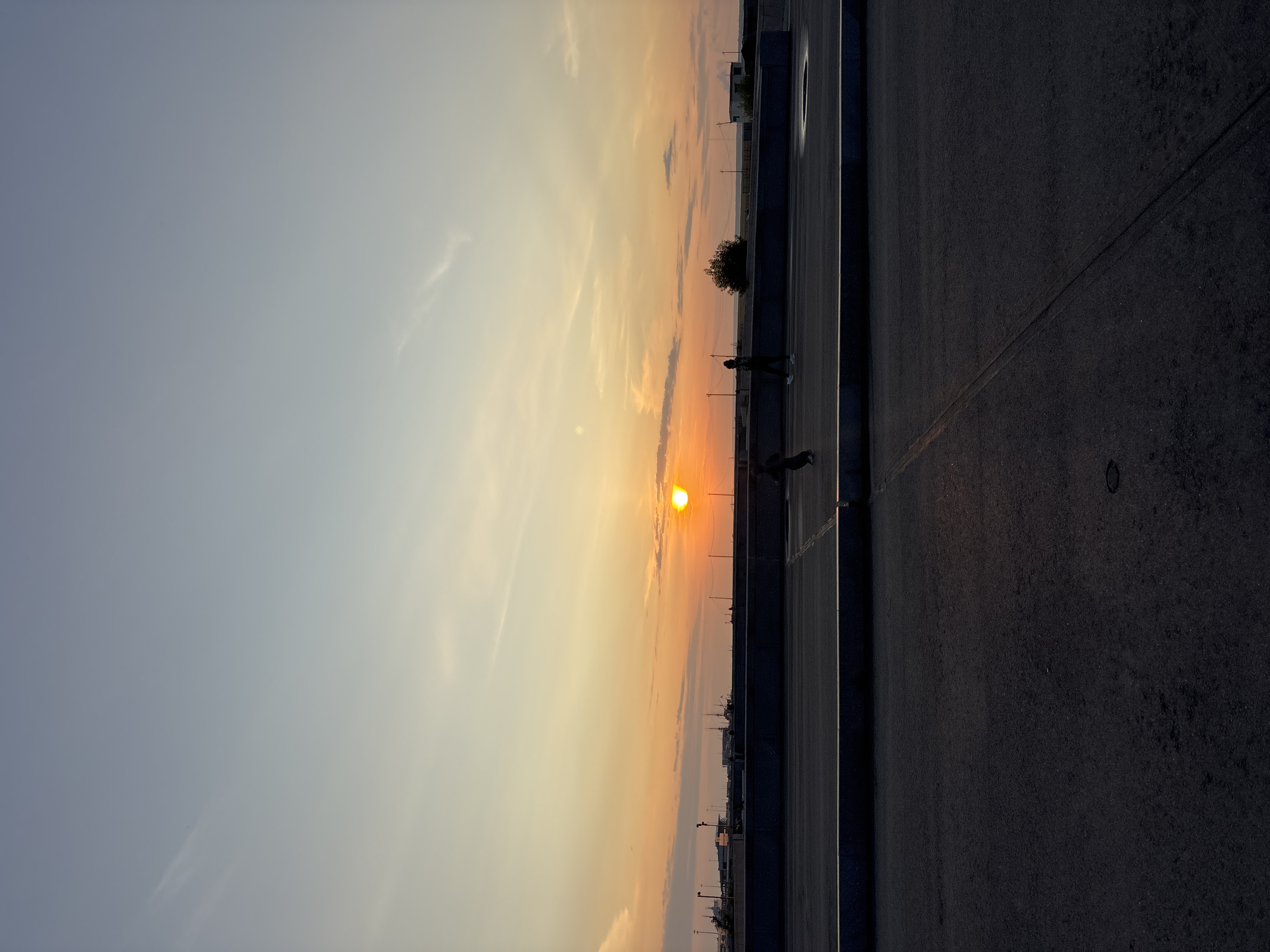 A sunset scene unfolds over the port of Saint Petersburg, Russia, captured from a high vantage point, likely a rooftop or elevated walkway. The sky is a gradient of soft blues and oranges, with the sun a vibrant disc low on the horizon. Silhouetted against the colorful sky is a long, dark structure, possibly a pier or a retaining wall, dotted with what appear to be ships or boats docked in the distance. Below this waterfront, a dark asphalt surface stretches into the foreground, suggesting a public space like a parking lot or promenade. Two figures are visible on the elevated walkway; one is a child in motion, and another adult stands nearby. The overall mood is serene, with the setting sun casting a warm glow over the urban landscape.