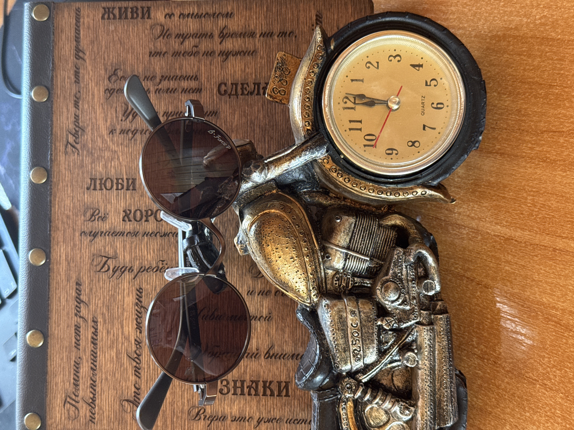This image features a close-up, overhead shot of several objects placed on a wooden surface. The primary focus is on a decorative clock with a steampunk aesthetic, designed to resemble a mechanical contraption. It has a round, gold-colored clock face with black numerals and hands, and is mounted on an intricate, metallic structure.
In front of the clock, a pair of round, dark-lensed sunglasses are perched, their metallic frames partially obscuring the clock's lower portion. The sunglasses appear to be the "B-Force P" brand.
The wooden surface itself is not smooth but rather engraved with Russian text, suggesting it might be a decorative plaque or a journal cover. The text is in a stylized, cursive font. Some visible phrases include "ЖИВИ смыслом" (Live with meaning), "Не трать время на то, что тебе не нужно" (Don't waste time on what you don't need), "ЛЮБИ" (Love), and "Будь ребё" (Be a child).
The background is a plain, textured wooden surface, likely a table or desk, with a slight sheen. The lighting appears to be from above, creating soft shadows. The scene is static, with no discernible activity taking place, and it conveys a sense of contemplation or appreciation for decorative and inspirational items. The overall impression is one of curated display, with a blend of functional objects and aesthetic elements.