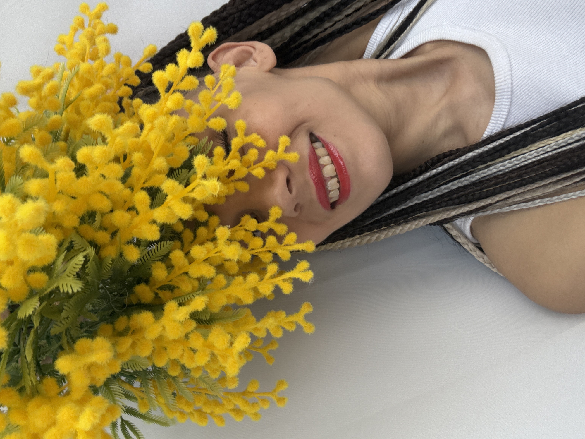 A person is lying on a white surface with a large bunch of yellow flowers partially obscuring their face. The person has brown and blonde braided hair and is wearing a white tank top. They are smiling with red lipstick. The location is unknown.