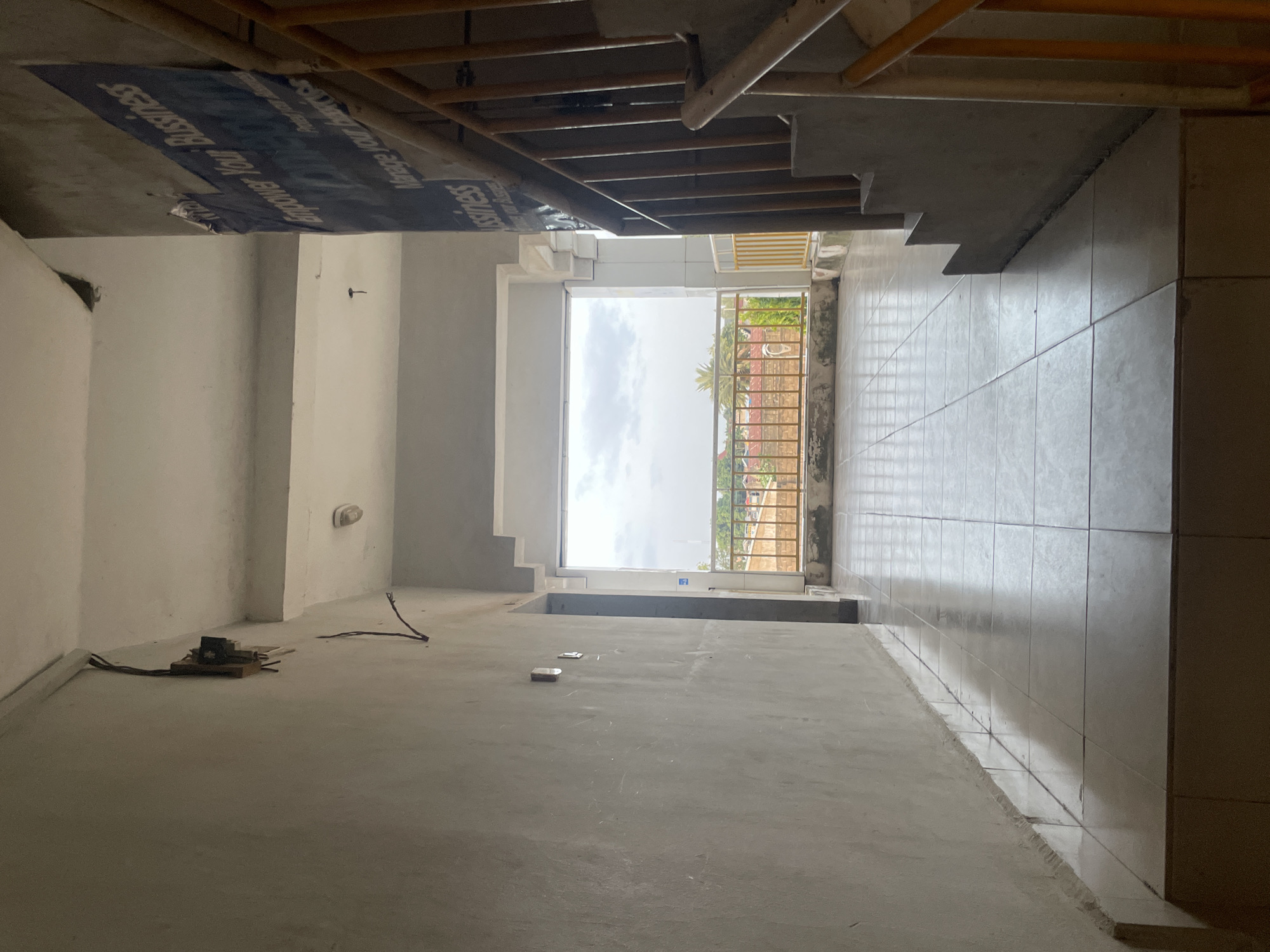 The image shows a narrow, tiled hallway in what appears to be a building under construction or in disrepair, located in Jalingo, Nigeria. The hallway is lit from the end by a large opening that looks out onto a cloudy sky and some greenery. To the right, a concrete staircase with a yellow railing leads upwards. The walls are bare concrete, with some exposed wires and electrical boxes visible on the left. A large blue banner with text is partially visible on the right wall, near the top of the stairs. The floor is tiled with large, light-colored square tiles that reflect the light. There are no people visible in the image. The overall impression is one of an unfinished or neglected indoor space with an outdoor view.