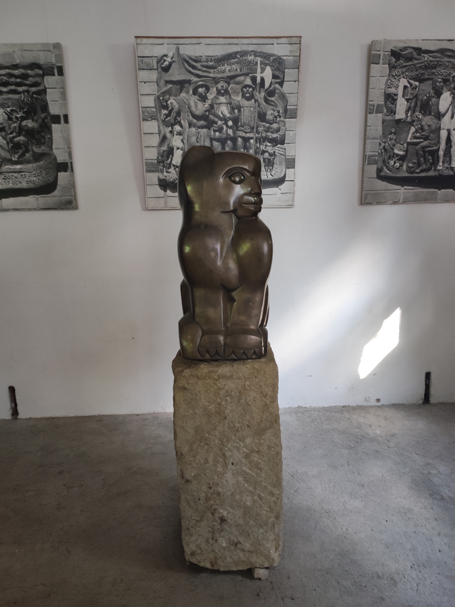 The image shows a bronze sculpture of a stylized human figure, possibly inspired by African art, displayed on a rough-hewn stone pedestal. The sculpture is dark brown with a smooth, polished finish. The figure is seated with its legs bent and hands resting on its knees, and its head is turned to the side, with prominent facial features including a large nose and mouth. The sculpture is positioned in the center of the frame, with a white wall as the background.
On the wall behind the sculpture, two framed artworks are visible. These artworks appear to be relief sculptures or bas-reliefs, rendered in black and white. The artwork on the left depicts a scene with multiple figures and what looks like water or waves. The artwork in the center features a group of figures, some holding weapons, and a banner. The artwork on the right also shows a scene with several figures in a domestic or communal setting. All three artworks have a textured, almost stone-like appearance.
The setting appears to be an indoor exhibition space, possibly a museum or gallery, given the display of art. The floor is a textured concrete or stone surface. Sunlight streams in from the right side of the frame, casting a bright, diagonal reflection on the white wall. The overall impression is one of artistic display, with the central sculpture being the primary focus. The style of the artworks and the sculpture suggests a possible exhibition related to history, culture, or social commentary. The location context is Dresden, Germany, but there are no explicit visual cues within the image to confirm this. The time of day is suggested by the sunlight, indicating it is daytime.