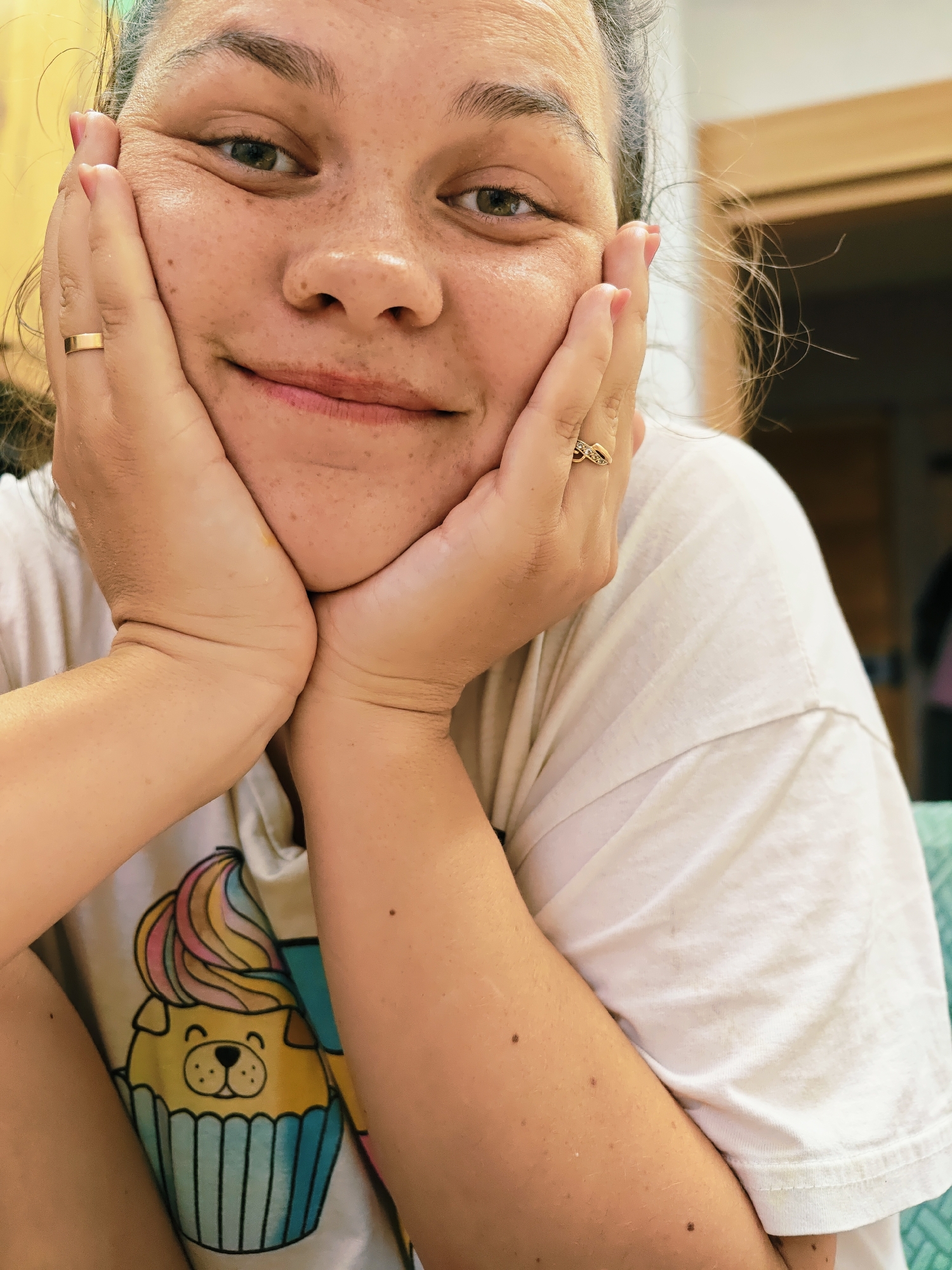 A medium, close-up shot depicts a woman with fair skin and freckles smiling slightly and resting her chin on her hands. She wears a plain white t-shirt with a colorful graphic of a dog inside a cupcake. She has a plain gold band on the ring finger of her left hand, and a more ornate ring on the ring finger of her right hand. Background details are minimal, showing a door frame in the distance. The location is the city of Chaikovsky, Russia.