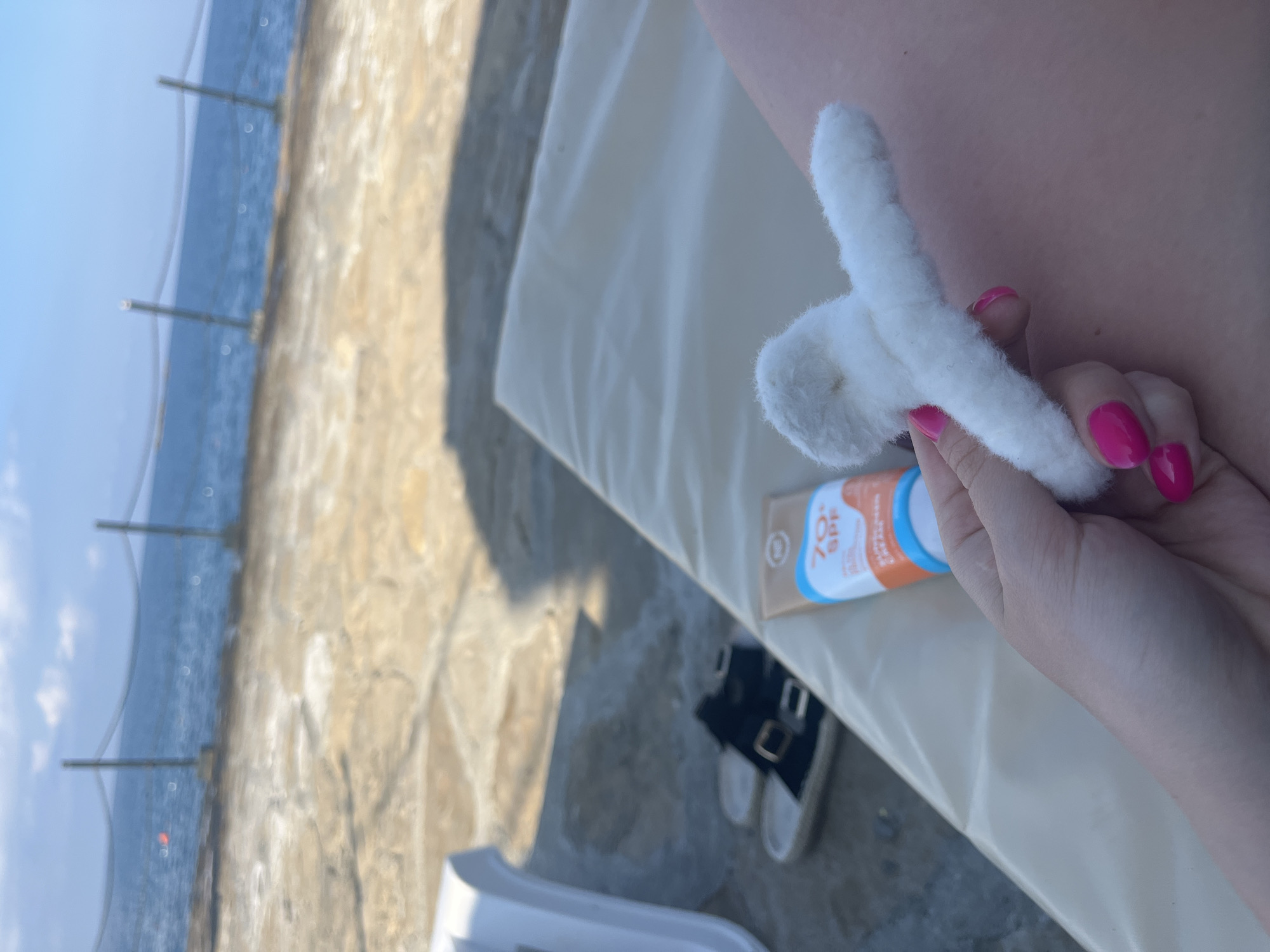 In Alanya, Turkey, a person's hand with bright pink fingernails holds a white fluffy object, possibly a hair accessory, against a backdrop of a sun lounger, a tube of sunscreen labeled "70 SPF," and a pair of black sandals. Beyond the lounger, the blue sea stretches out, bordered by a stone pathway and a fence, suggesting an outdoor setting like a beach or poolside area. The overall scene evokes a sense of relaxation and preparation for outdoor activities.