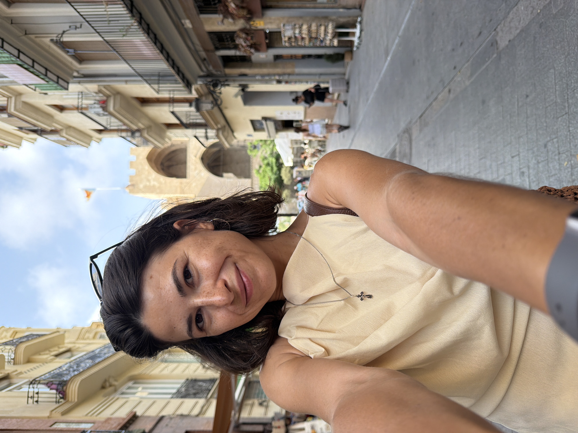 A woman is taking a selfie in a European-style city street. The woman has dark hair and is wearing a light-colored dress and a silver necklace with a cross. She is smiling and looking at the camera. The background of the image shows a narrow street with buildings on either side. The buildings are several stories high and have balconies. There is a flag flying from a building in the distance. There are a few other people walking in the background, and some outdoor seating for a cafe. The sky is blue with some clouds.