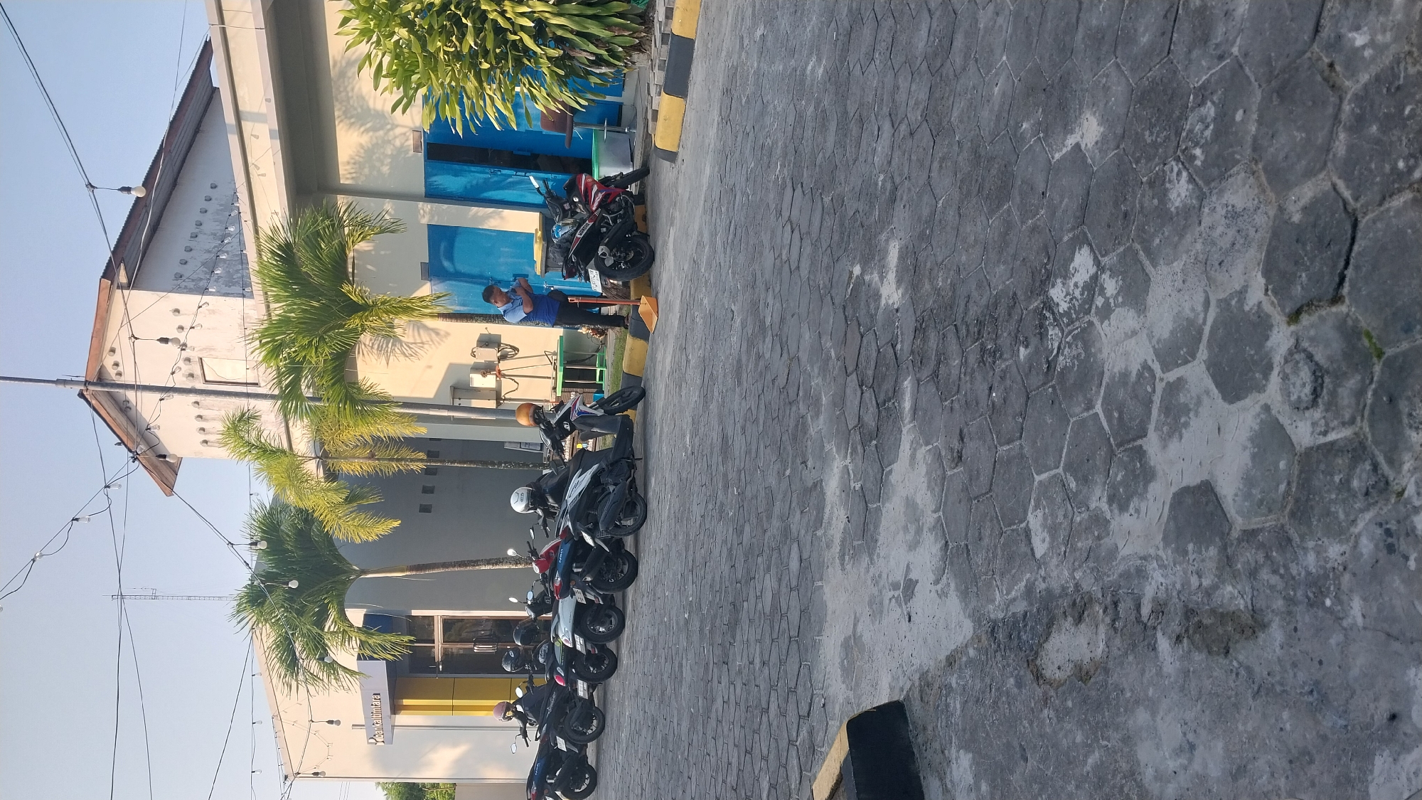 A man in a blue shirt and dark pants is sweeping the pavement in front of a building. He is holding a broom in his right hand and a dustpan in his left. There are several motorcycles parked in front of the building, and a palm tree stands to the left of the man. The building appears to be a bank, with a sign that reads "Bank Kaltimtara". The pavement is made of hexagonal interlocking stones, and there is a yellow and black curb. The sky is clear and blue. The scene is set in Penajam, Indonesia.