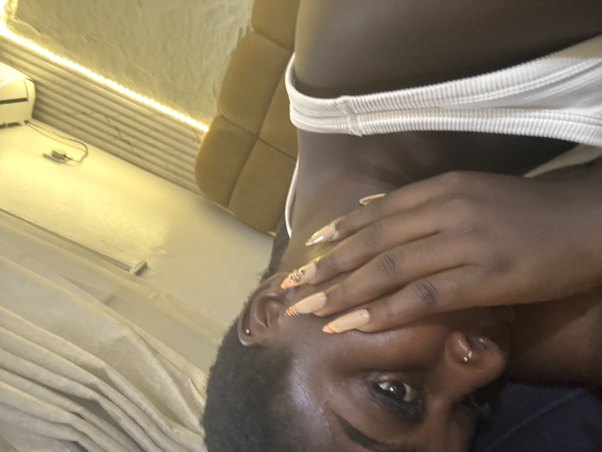 The image is a close-up, low-angle shot of a Black woman in what appears to be a bedroom in Abuja, Nigeria. She is lying down, with her head tilted and her hand covering part of her face. Her nails are long and decorated with a patterned design and small rhinestones. She is wearing a white tank top. In the background, there is a textured yellow wall with what appears to be a shelf and an air conditioning unit. There are also curtains and a padded headboard visible. A string of yellow LED lights runs along the wall. The lighting is warm and artificial.