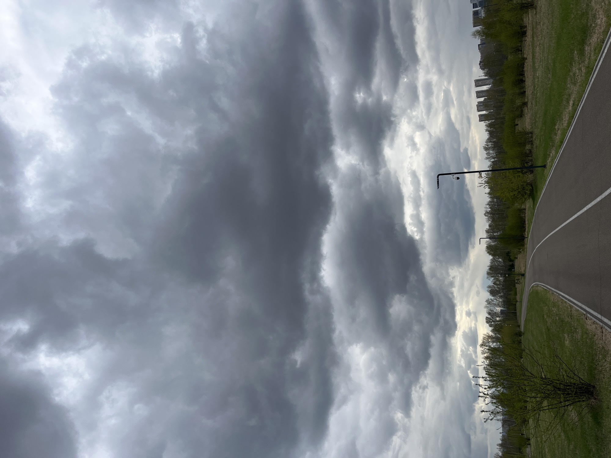 The image shows an outdoor scene under a cloudy sky. A paved path with a white dividing line runs along the right side of the frame, bordered by grass. A row of trees lines the edge of the path, interspersed with lampposts. In the distance, buildings are visible, suggesting an urban environment. The clouds are heavy and gray, creating a somewhat ominous atmosphere. Based on the location details provided, this scene is likely within the city of Krasnoyarsk, Russia.