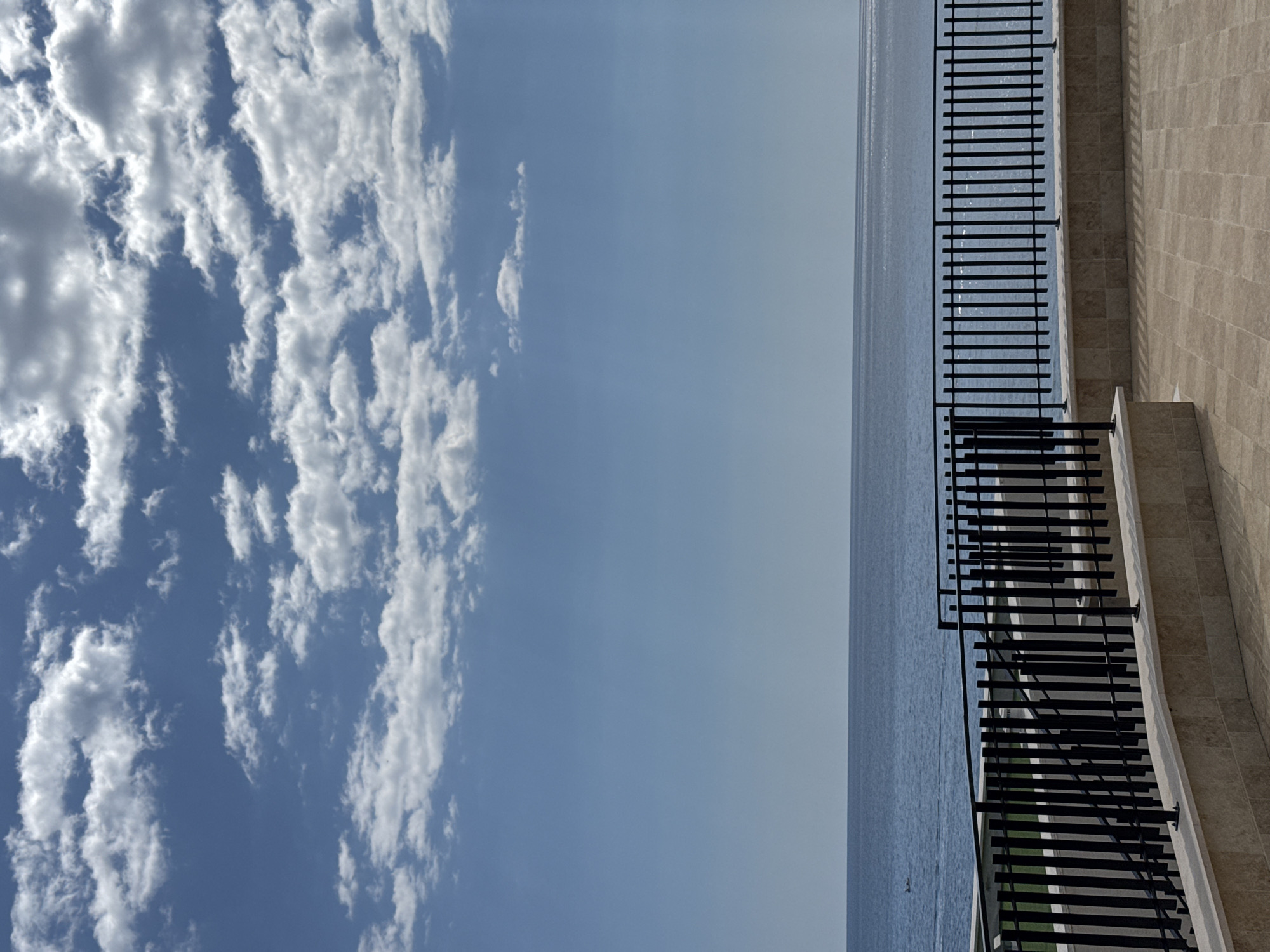 From Boreti, Montenegro, a view oriented vertically, the left side showing cumulus clouds in a blue sky. To the right, a body of water, possibly the Adriatic Sea, connects to a paved terrace featuring a wrought iron railing along the edge, with a light beige tiled wall as a backdrop.