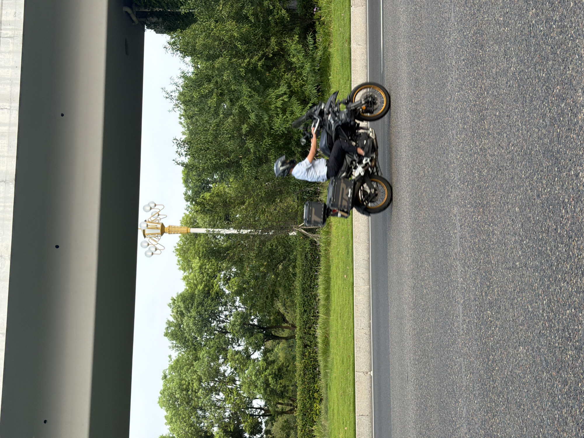 A person wearing a helmet rides a black motorcycle with saddlebags on a paved road. The motorcycle has gold-colored rims. The rider is wearing a light blue shirt and dark pants. The road is bordered by a grassy area with a hedge and lush green trees. In the background, a decorative lamppost with multiple lights is visible, and a concrete structure, possibly an overpass, runs across the top of the frame. The scene appears to be in the city of Jiangjin, China, suggested by the overall urban or suburban landscape.